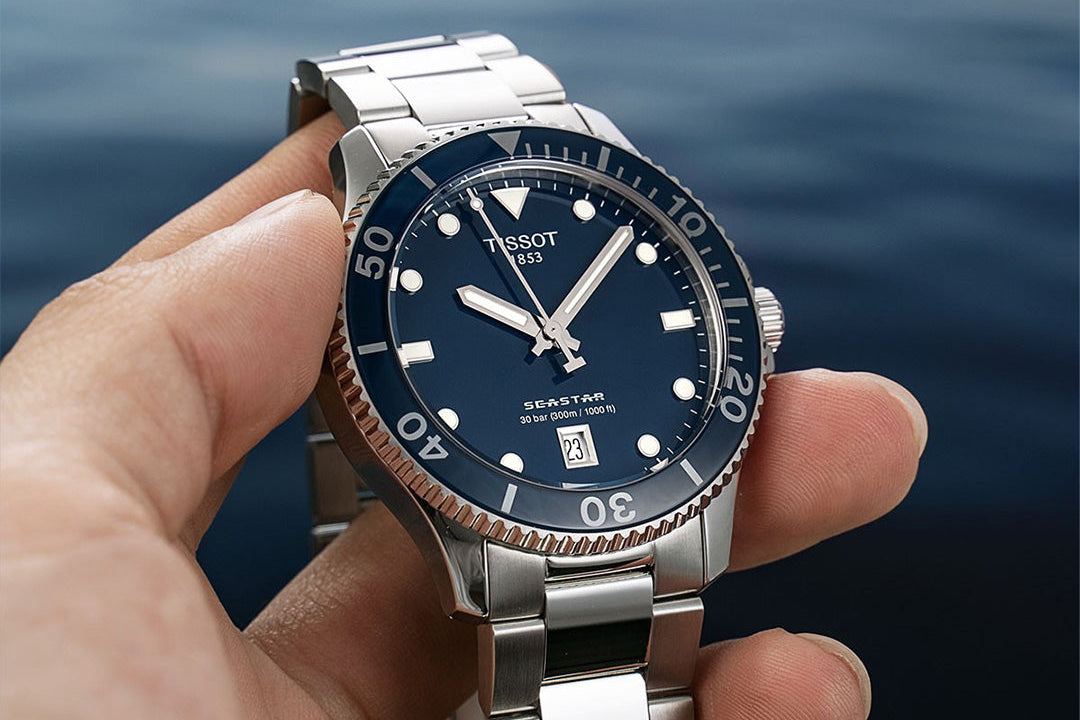 Silver watch with a blue dial held in a hand against a blurred water background