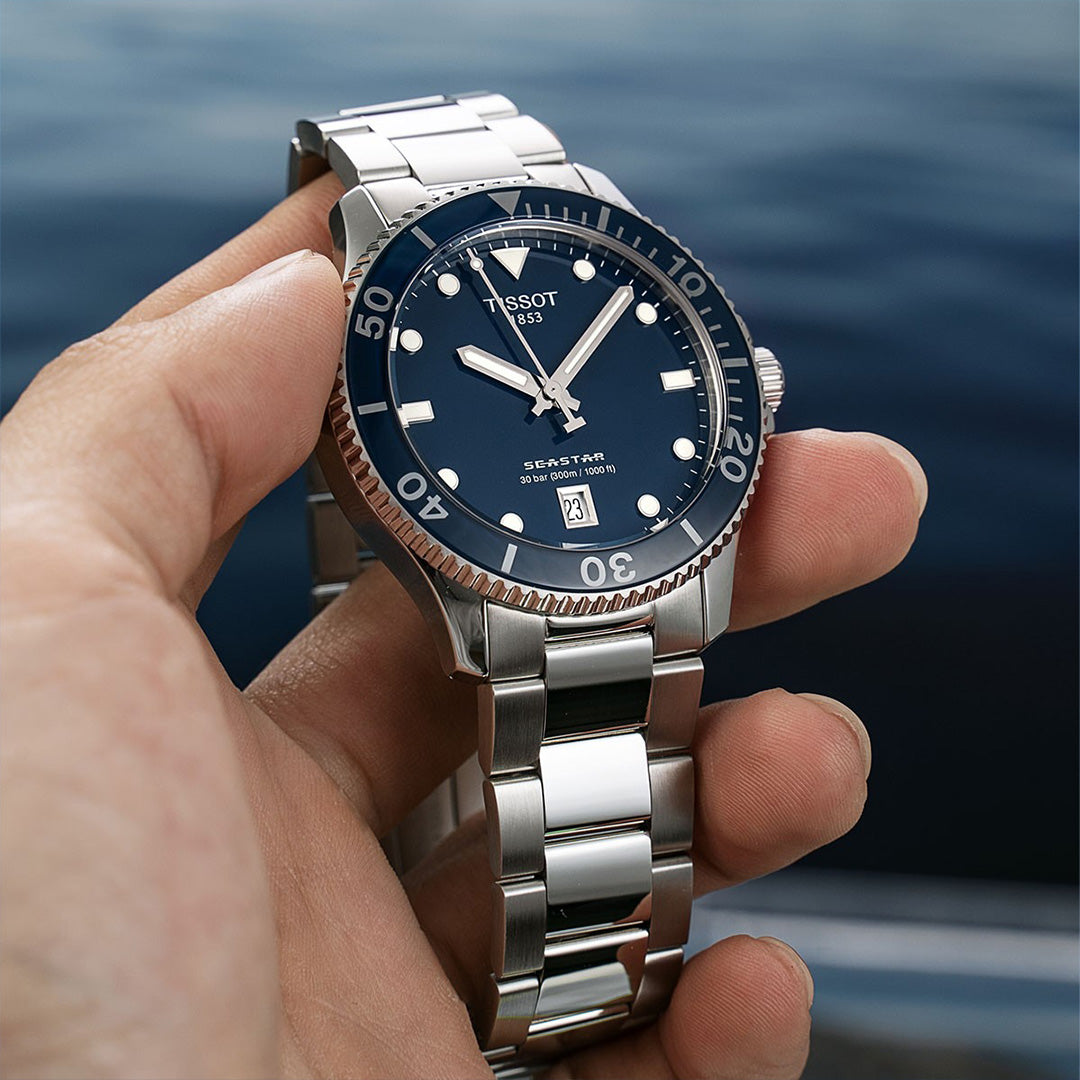 Silver watch with a blue dial held in a hand against a blurred water background