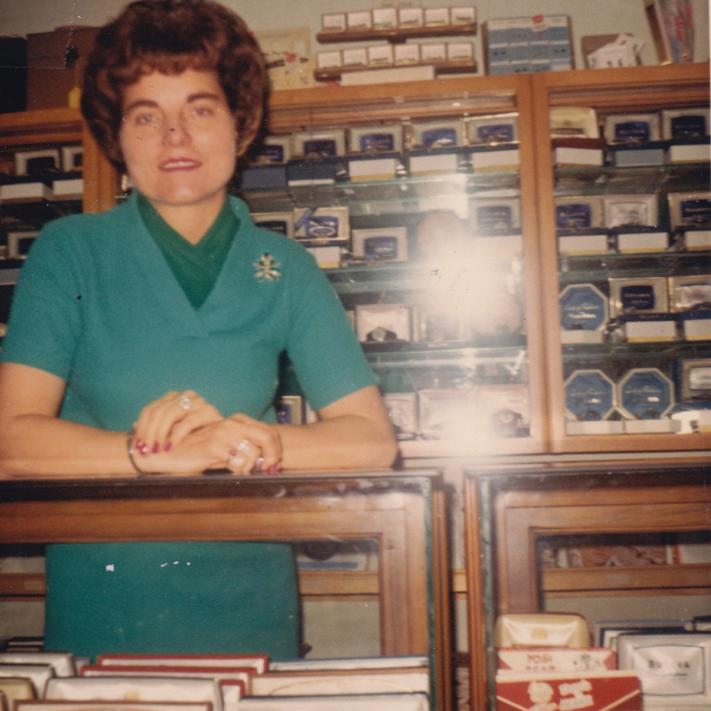 Woman in a teal shirt standing behind a counter with shelves filled with products in the background