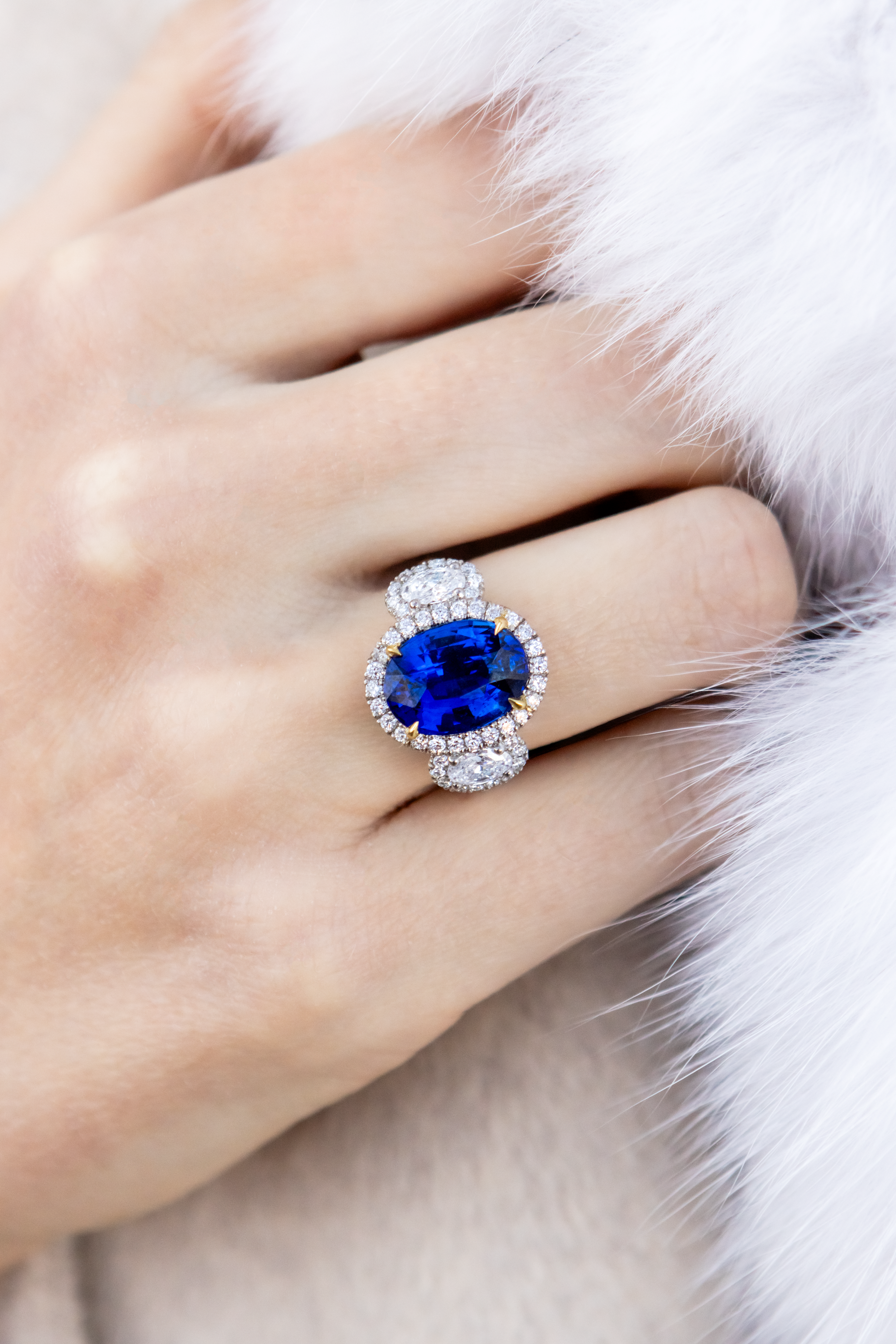 Close-up of a hand wearing a ring with a blue gemstone and diamond accents on a fur surface.
