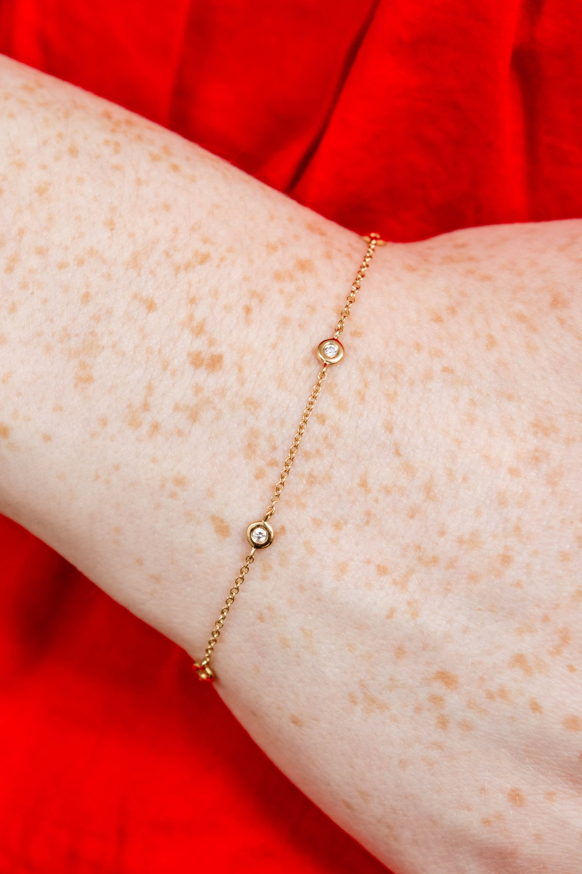 A delicate gold chain bracelet with two small bezel-set round clear stones, shown worn on a person's wrist against a vibrant red fabric background.