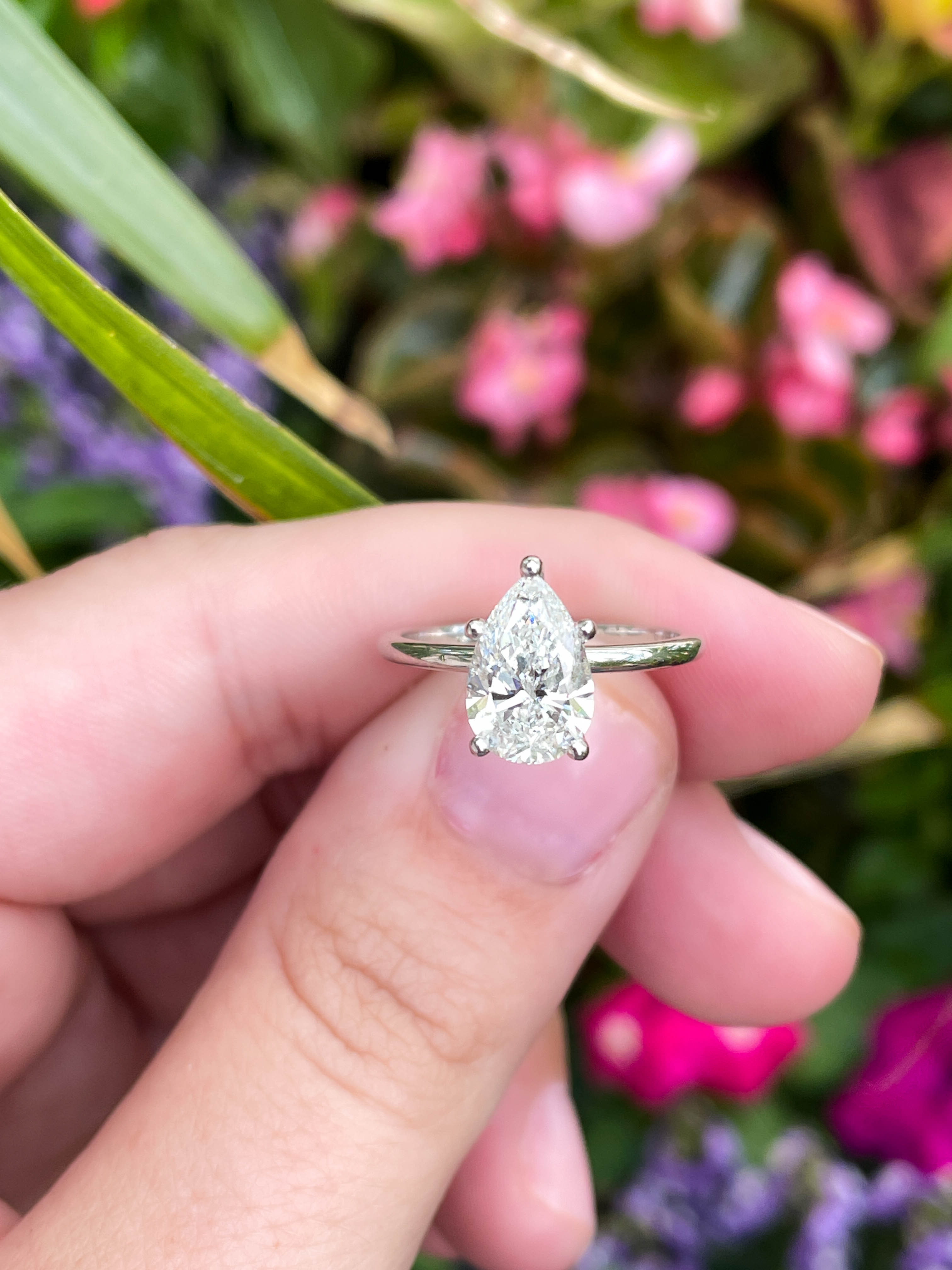 hand holding pear diamond silver ring in front of pink and purple flowers