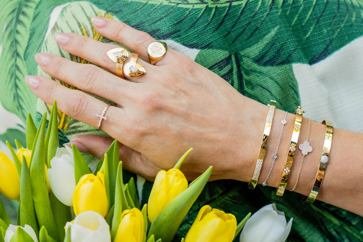 A close-up of a person's hand wearing multiple gold rings and bracelets, resting on a green tropical leaf-patterned fabric with yellow and white tulips in the foreground.