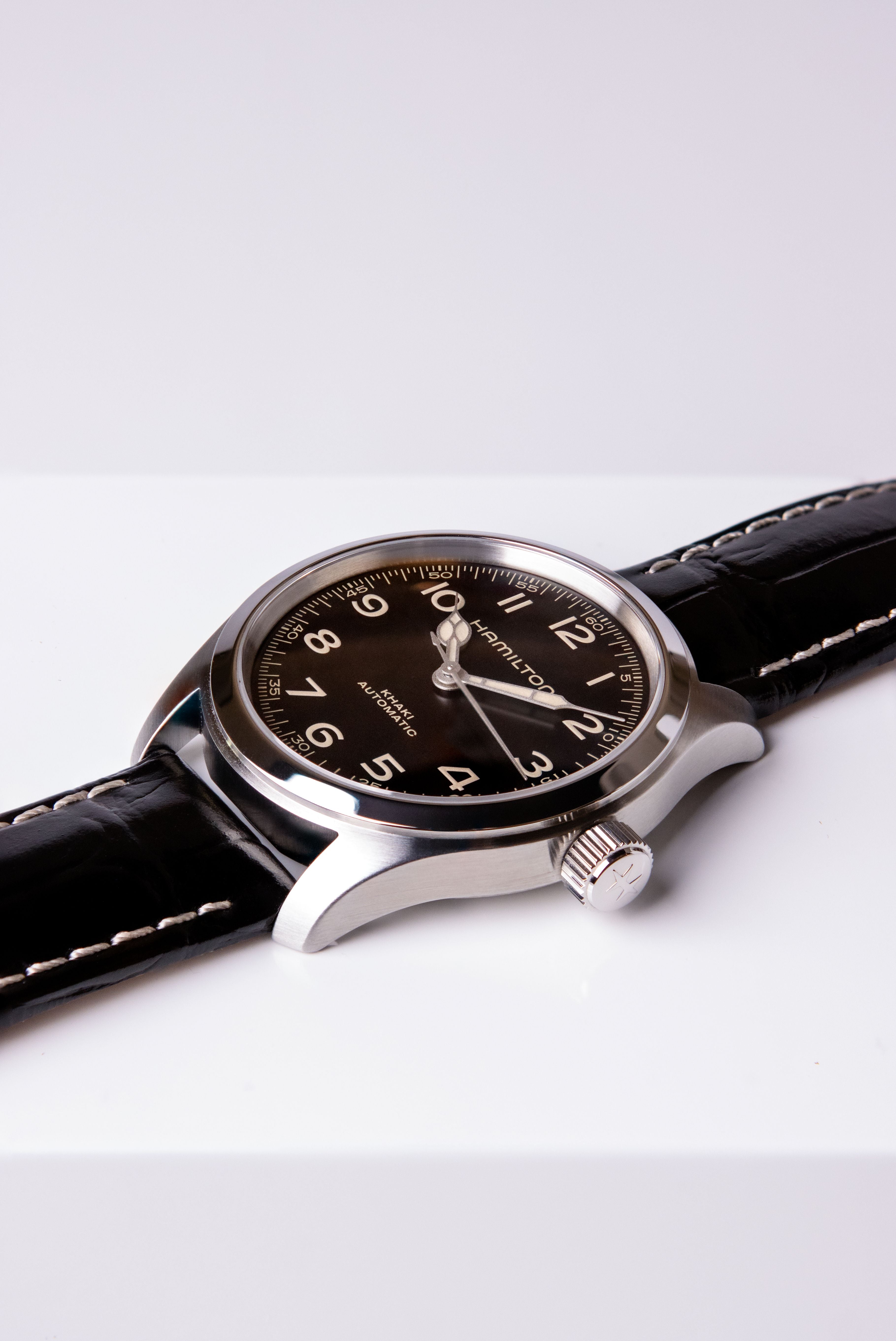 A Hamilton watch with a black dial, silver-tone stainless steel case, and a black leather strap with white stitching, displayed diagonally on a clean white surface.