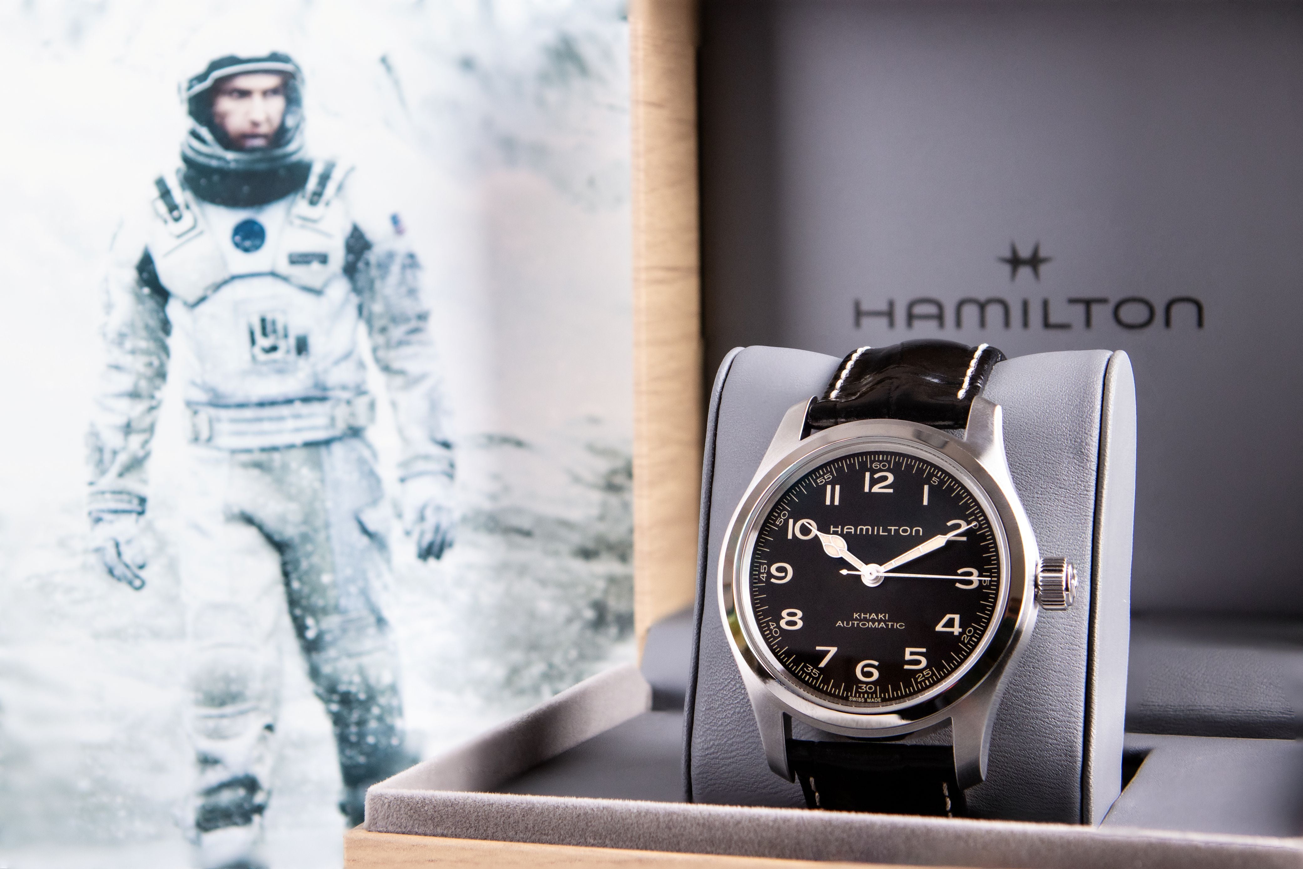 Hamilton watch displayed in a presentation box, featuring a black dial, silver-toned case, and black leather strap, with a Hamilton-branded box behind it and a poster of an astronaut to the left.