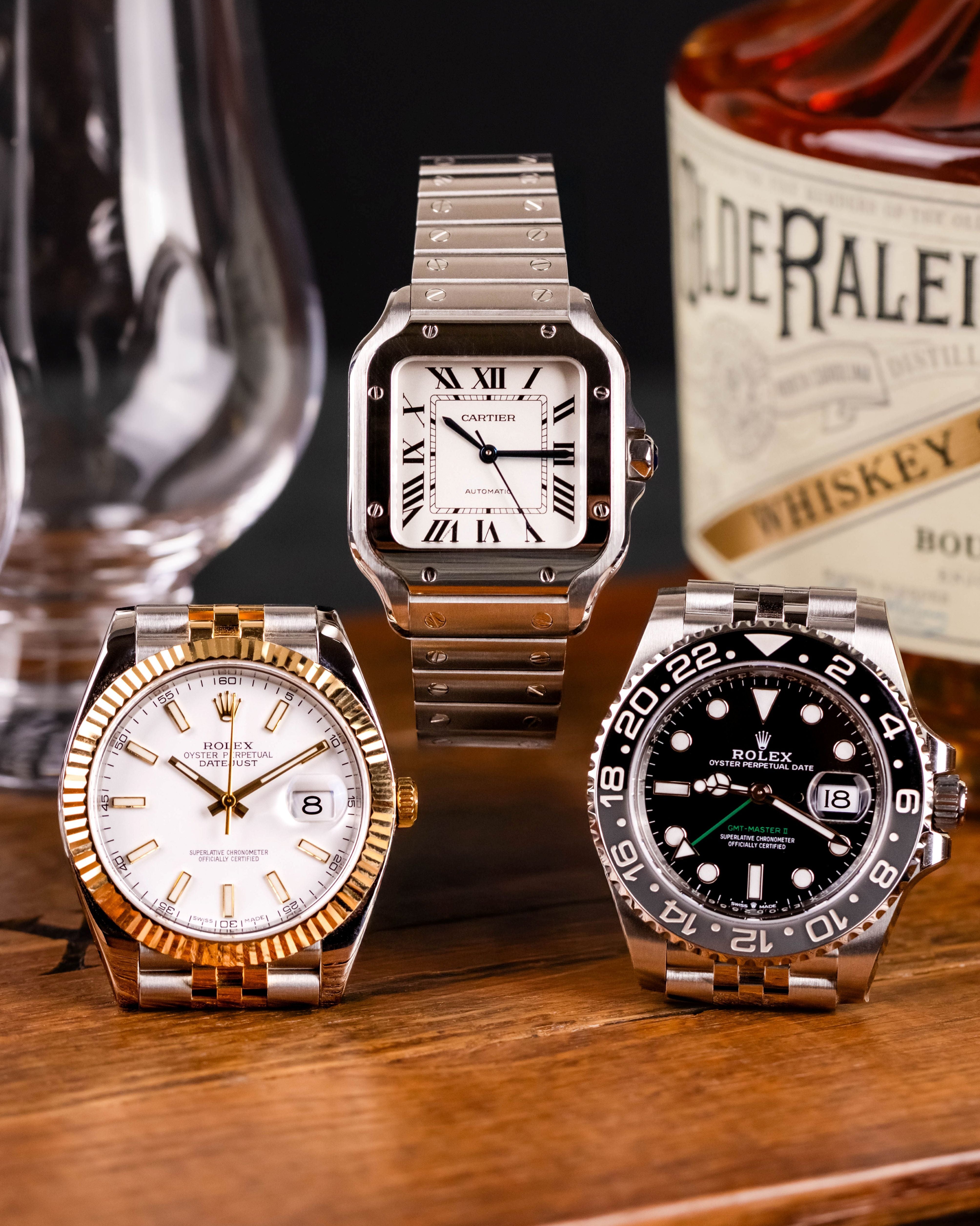 Three luxury watches on a wooden surface with a bottle of Rye Whiskey in the background.