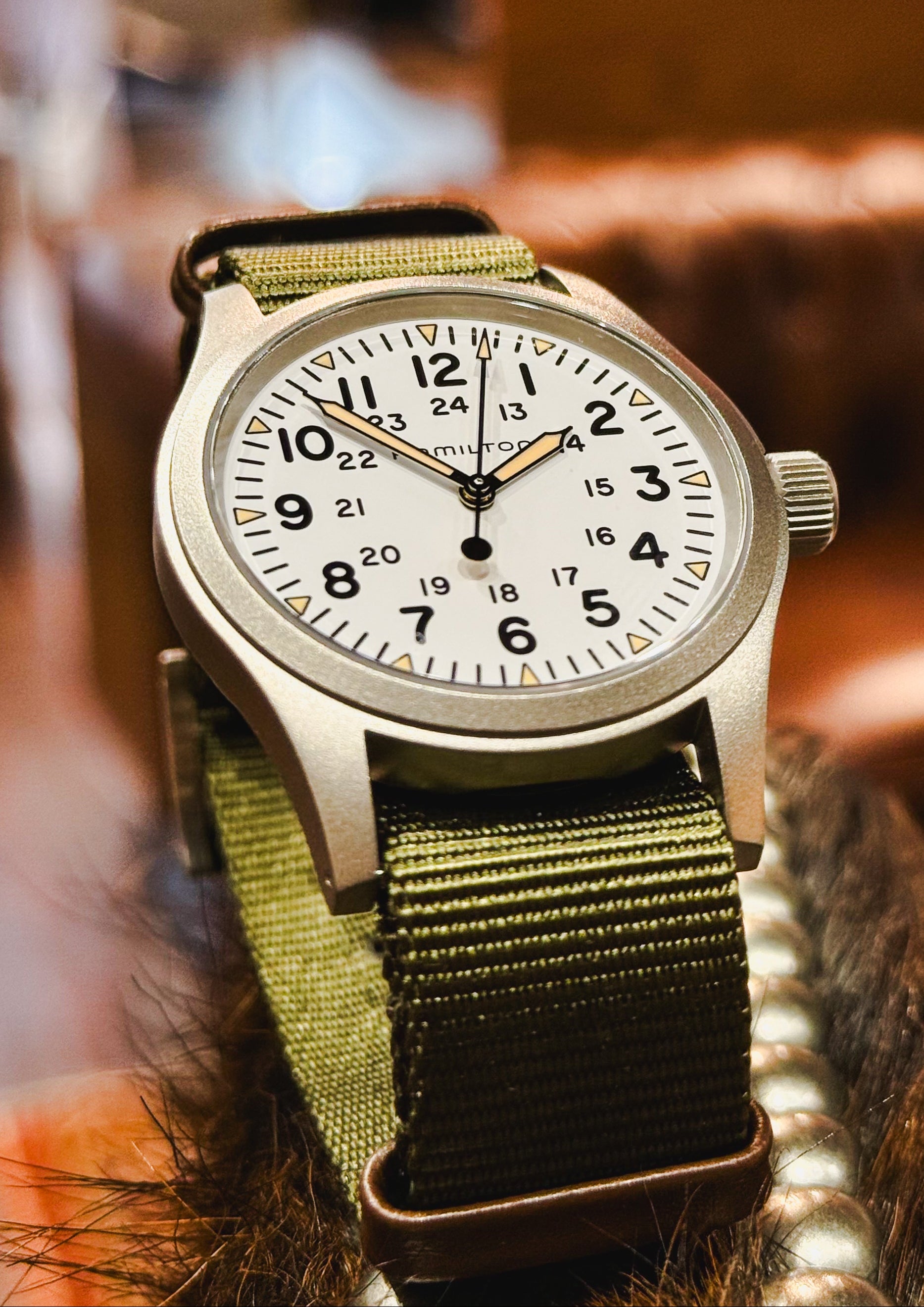 A close-up of a wristwatch with a silver-toned metal case, white dial with black Arabic numerals, and an olive green fabric strap, resting against a warm brown textured background.