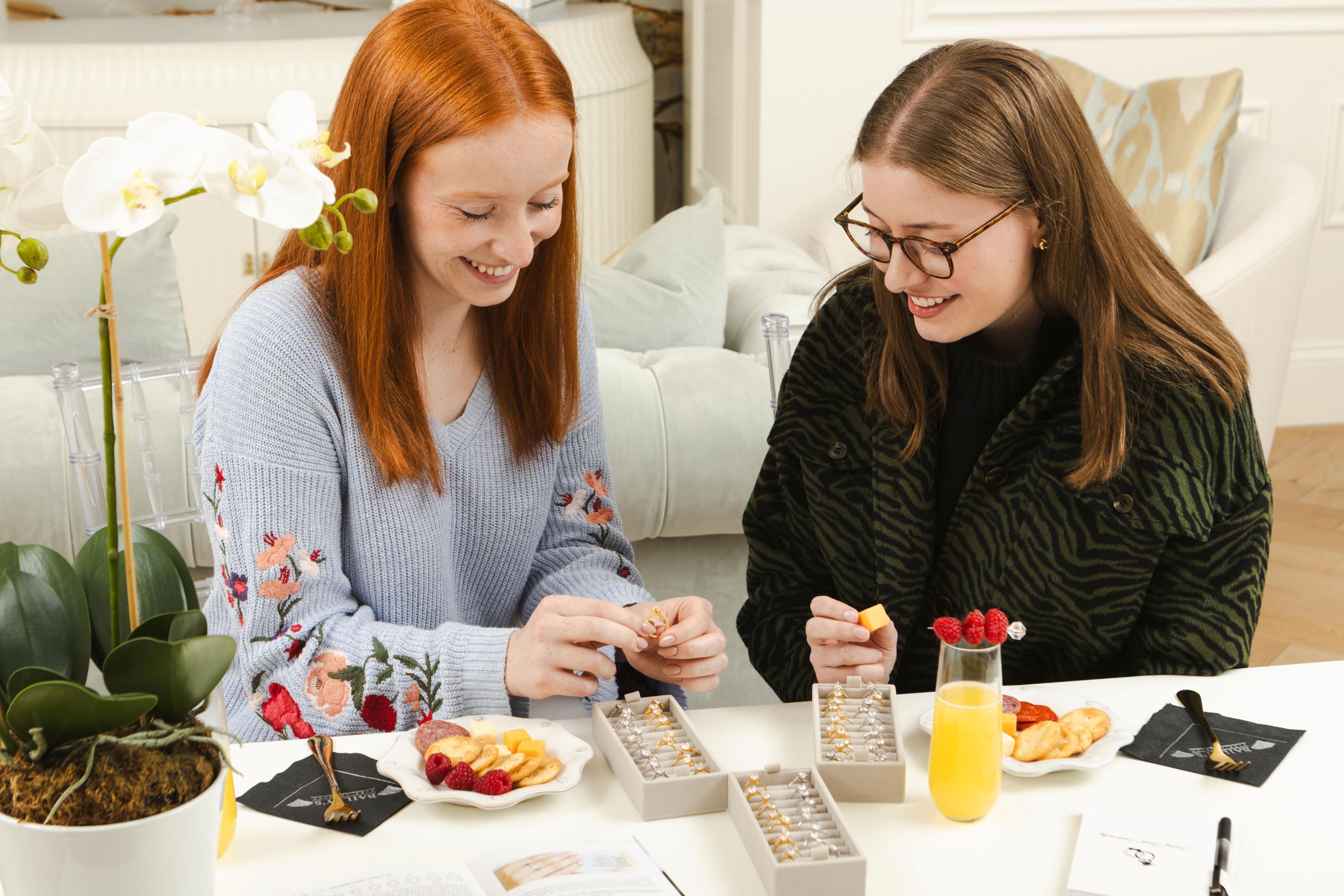 Two women sitting at a table with jewelry boxes and snacks, smiling and looking at jewelry.