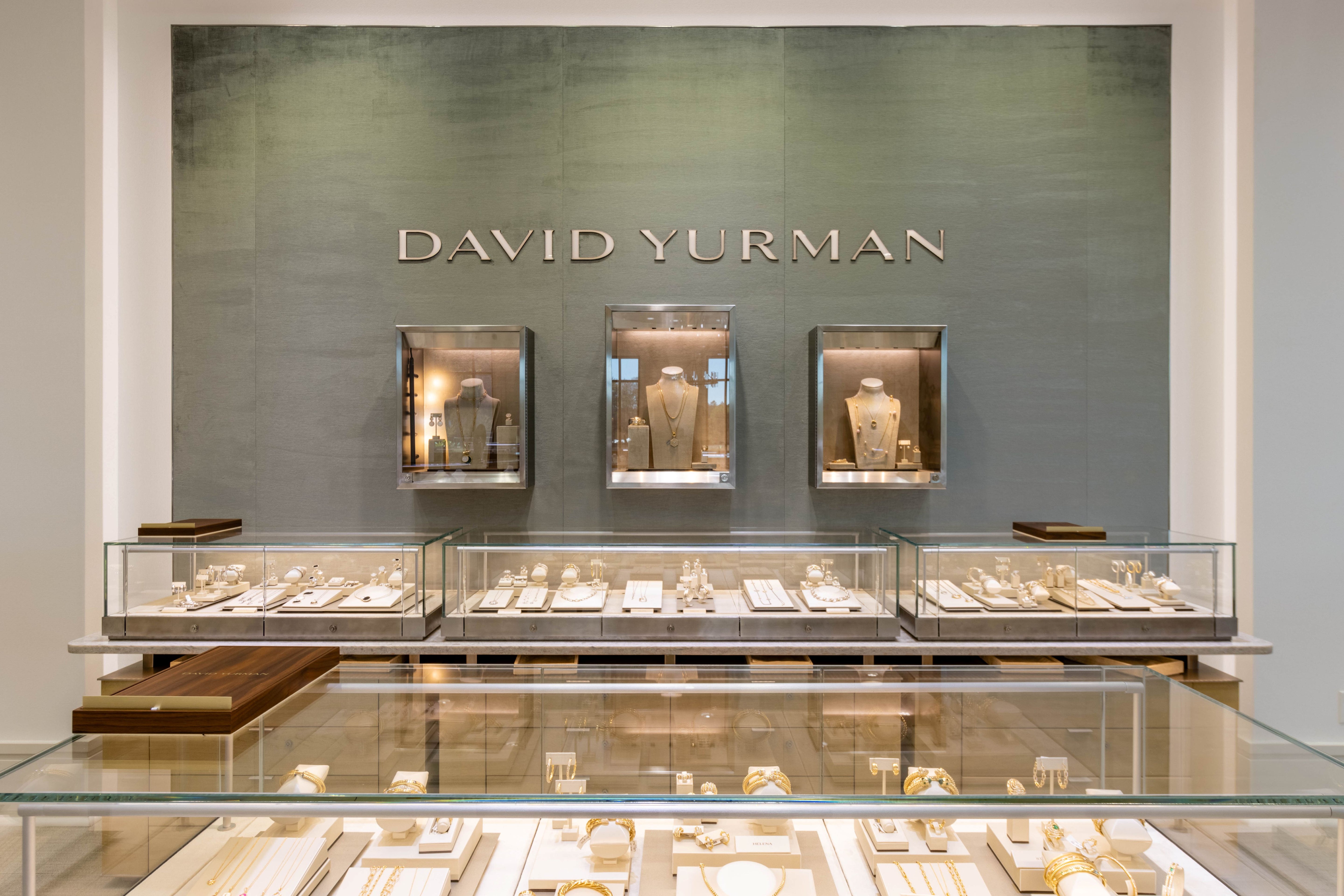 David Yurman jewelry display with various pieces on a glass shelf against a branded wall.