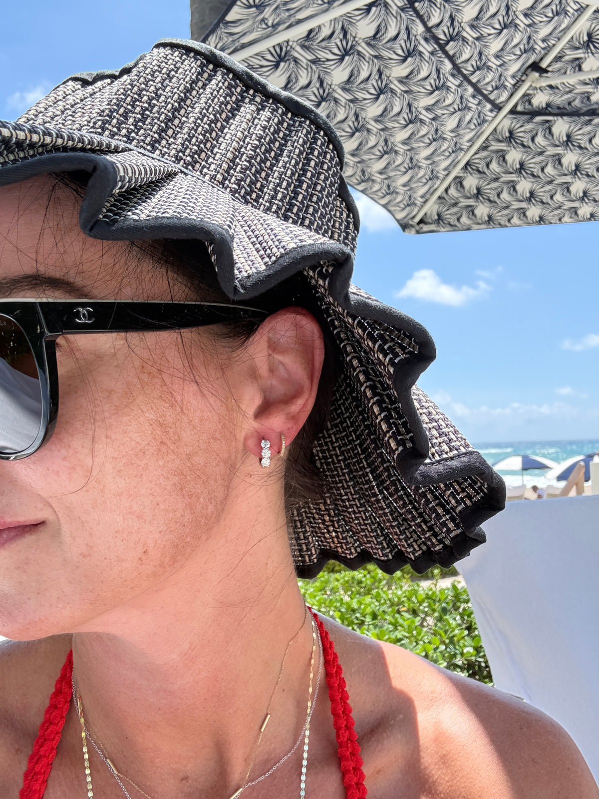 A close-up lifestyle shot of a woman wearing a black and white textured woven sun hat with a ruffled brim, black sunglasses, and a red braided top strap, outdoors under a patterned umbrella with a beach background.