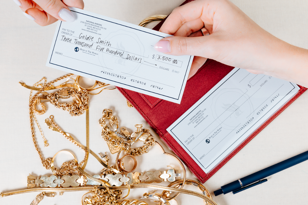 Two hands holding a check over a pile of gold jewelry on a light surface.