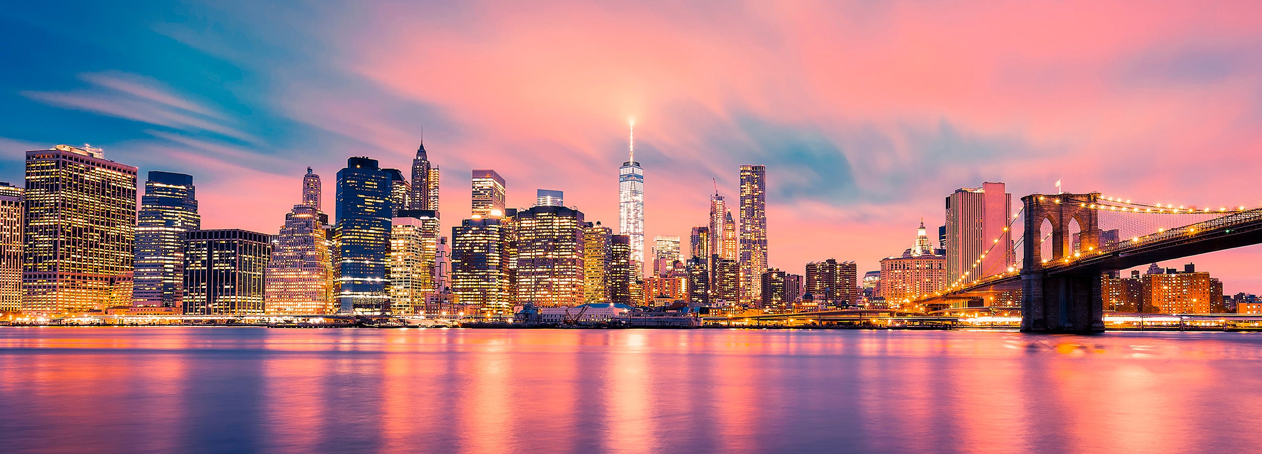 City skyline with illuminated buildings and a bridge at sunset.