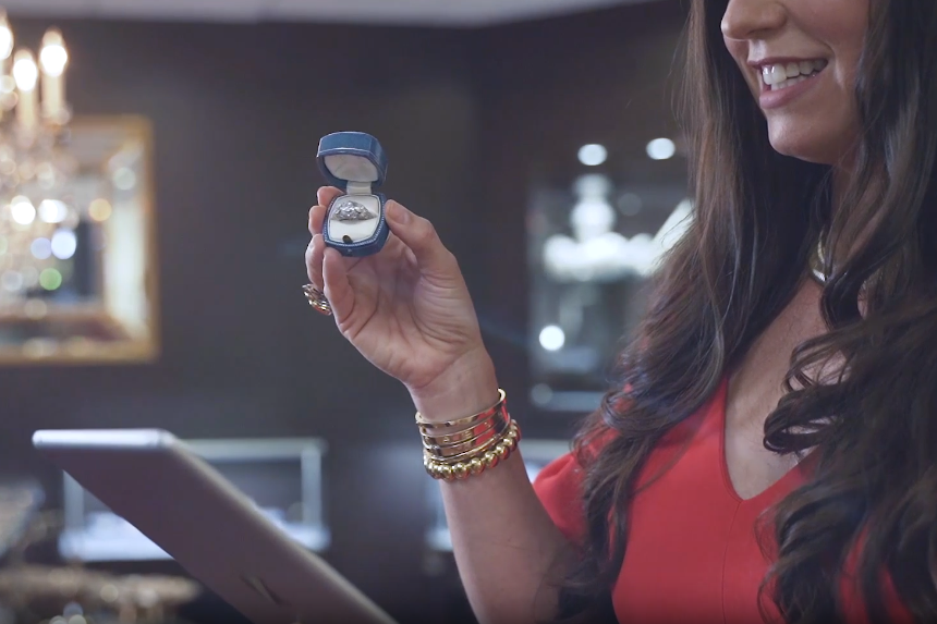 Woman holding a small jewelry box with a ring inside, smiling in a dimly lit room.
