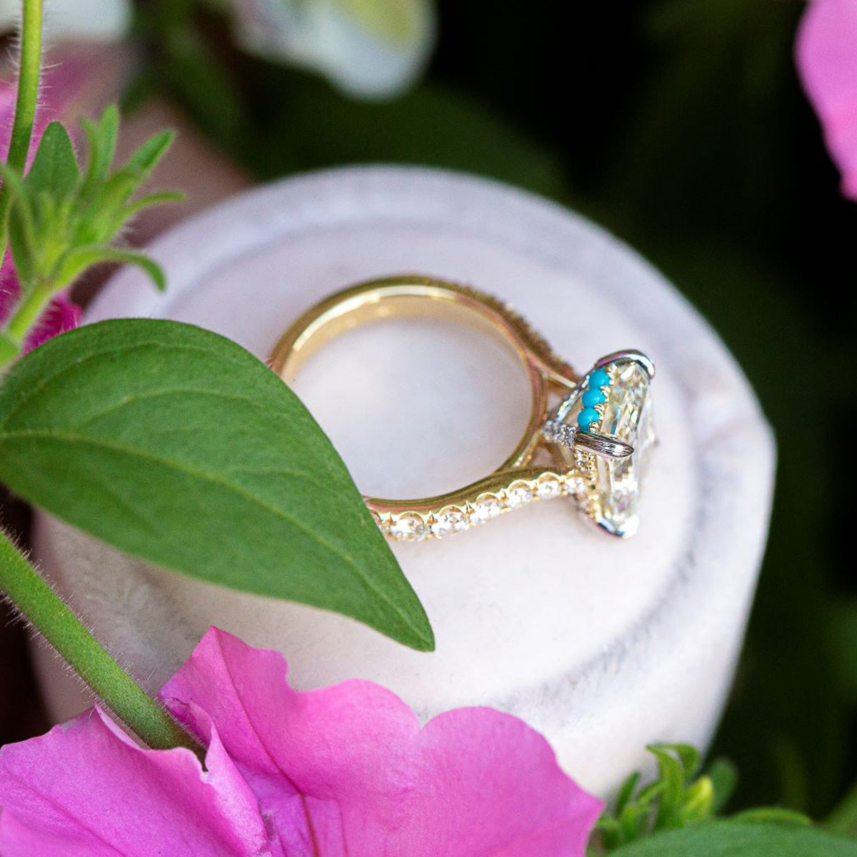 Gold ring with a diamond and blue stone on a white cushion, surrounded by pink flowers and green leaves.