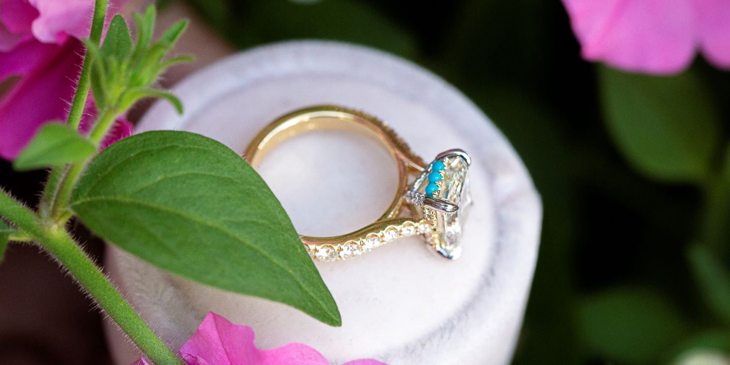 Gold ring with a diamond and turquoise stone on a white cushion, surrounded by green leaves and pink flowers.