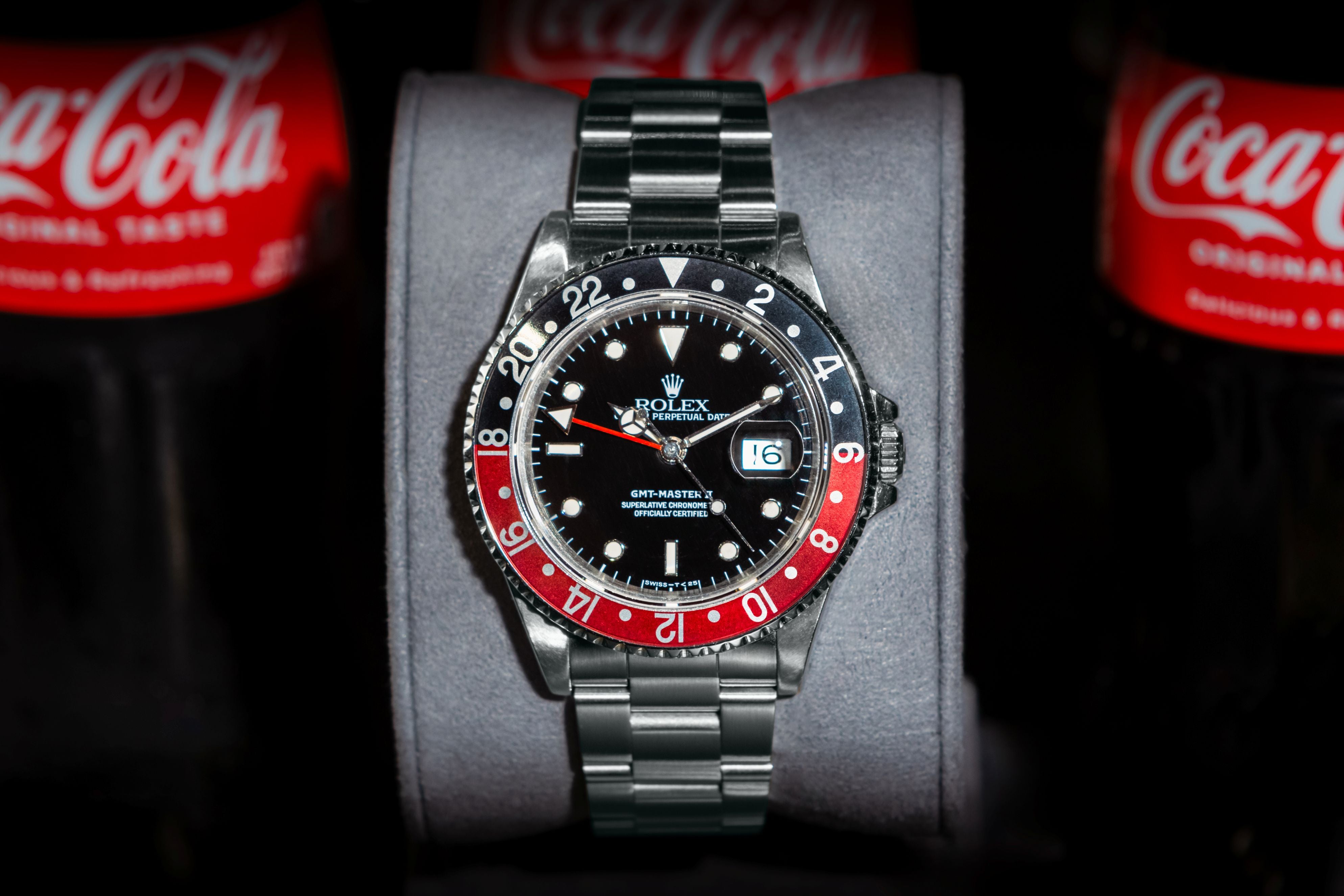 Rolex watch with red and black bezel on a gray stand against Coca-Cola bottles.