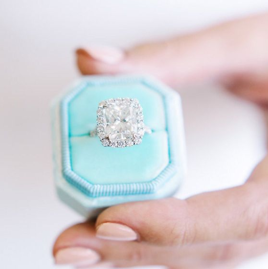 Diamond ring in a light blue hexagonal box held by a hand on a white background