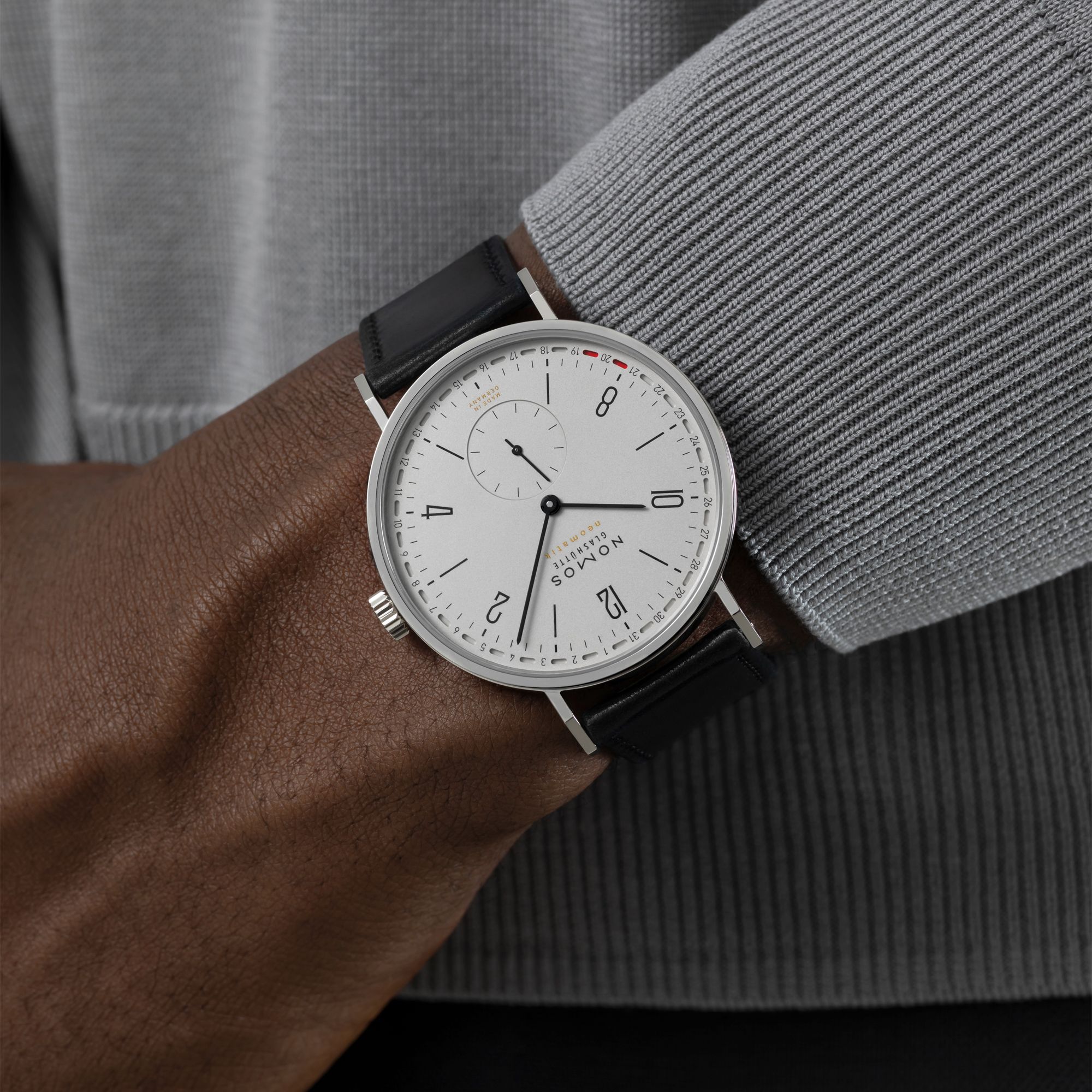 A close-up of a wrist wearing a London Watch silver-tone watch with a white dial and black leather strap, set against a gray ribbed sweater.