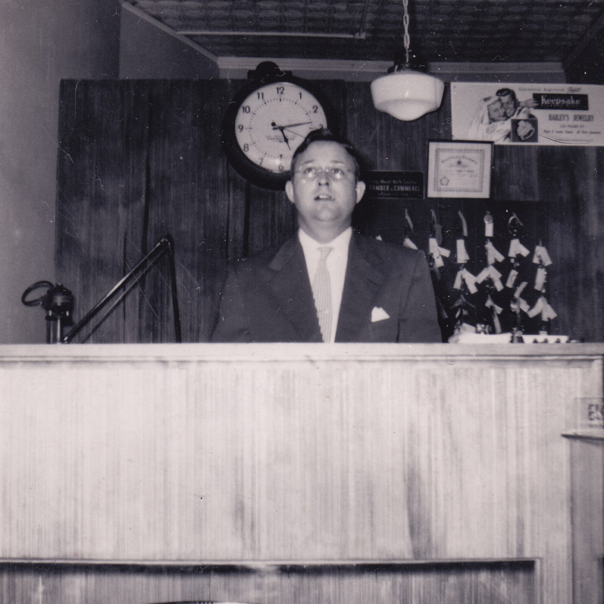 Man standing behind a counter with a clock and various items on the wall in the background