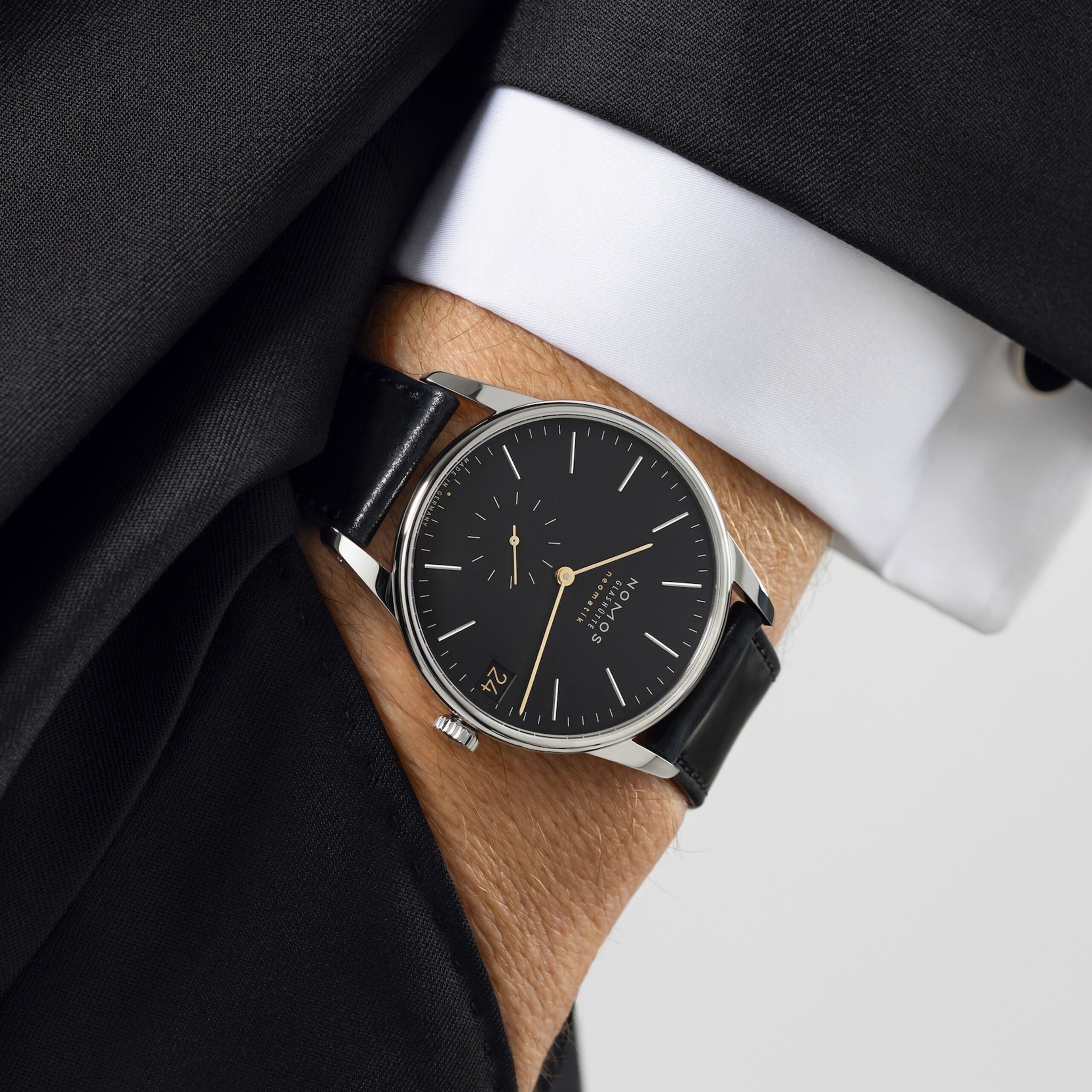 A close-up of a wristwatch with a black dial and black leather strap, worn on a person's wrist. The watch has a silver-toned case and gold-tone hands.