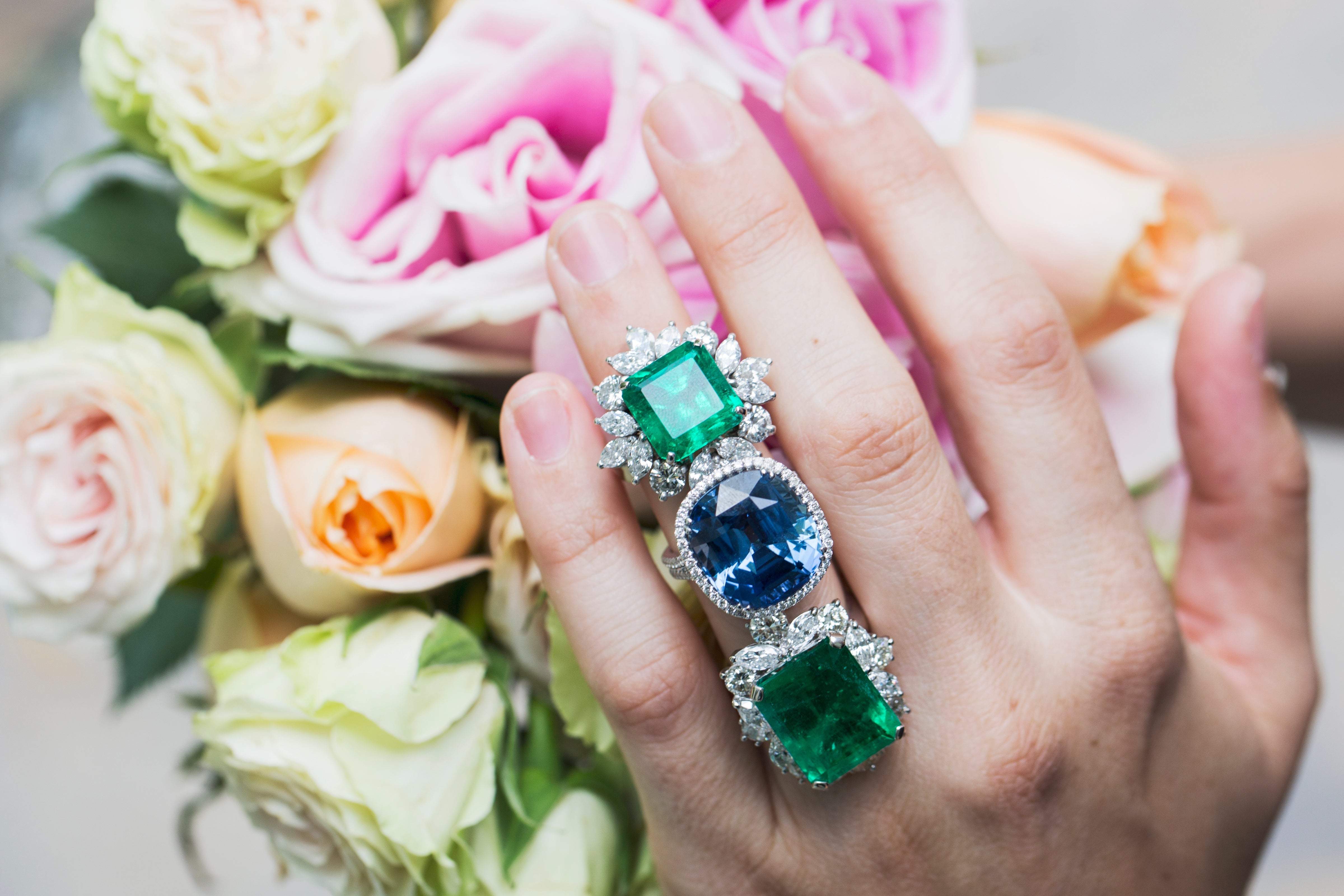 Hand wearing rings with emerald and sapphire stones in front of a bouquet of flowers