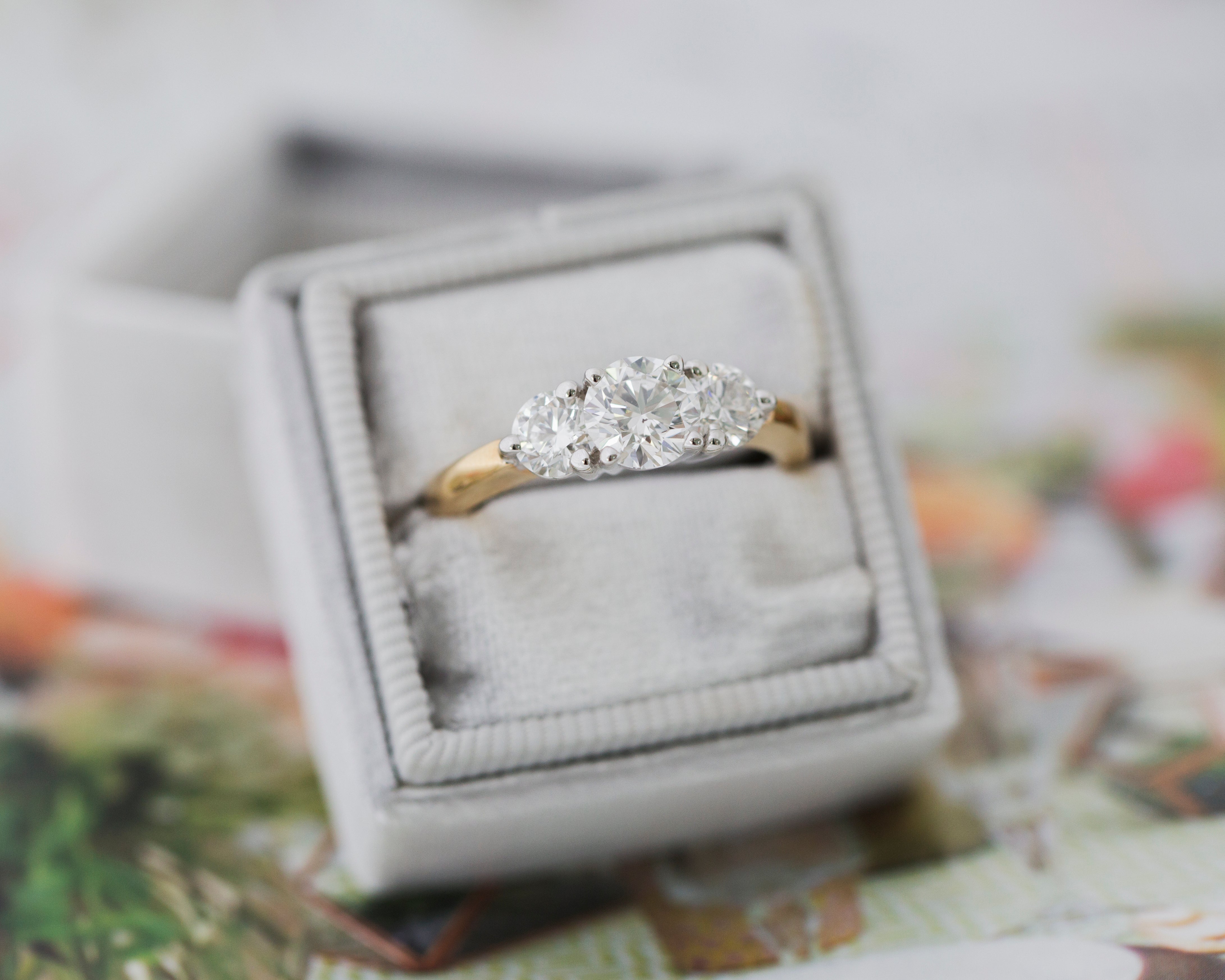 three stone diamond gold ring in a light grey box on a multicolored blurred surface