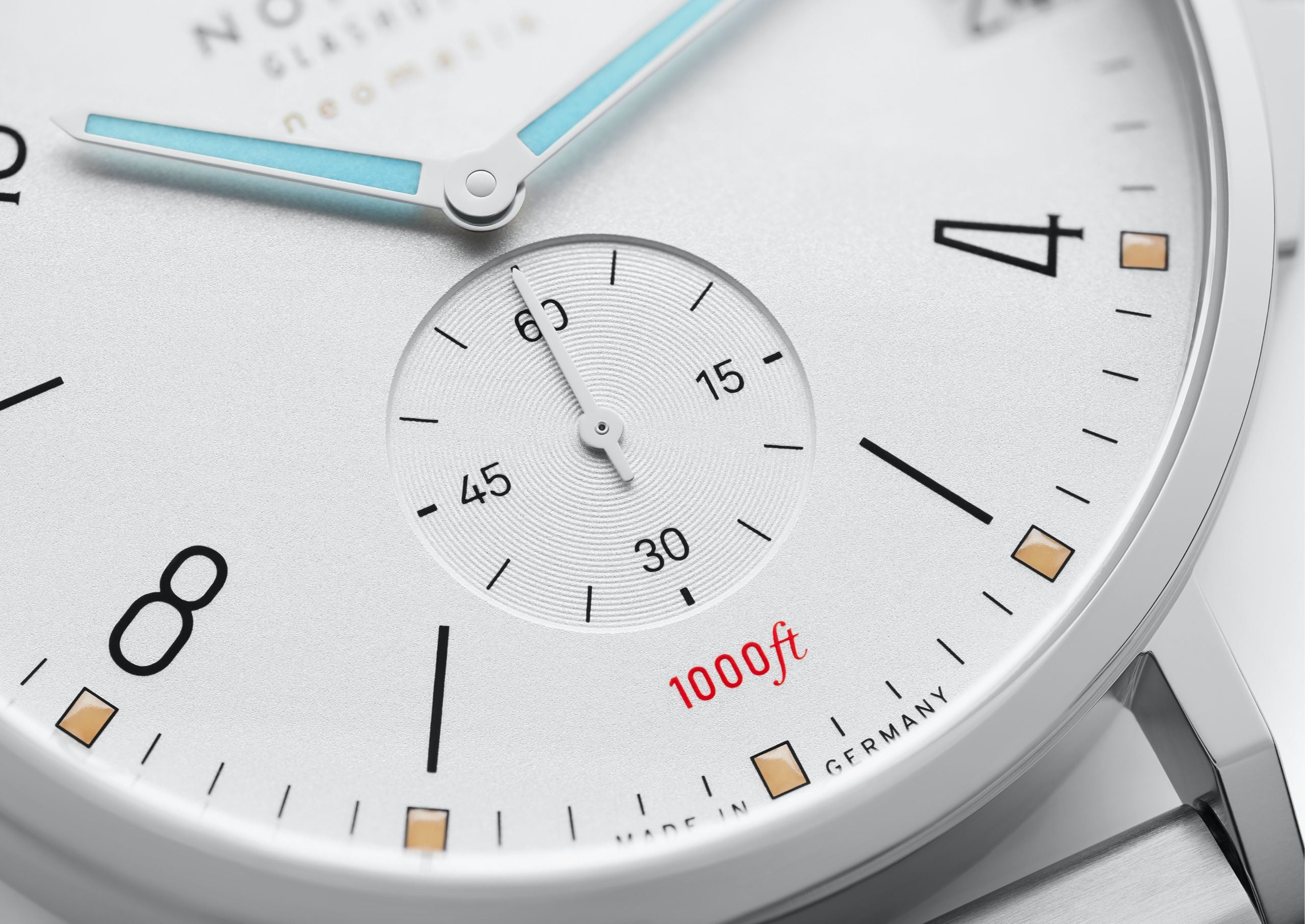 Close-up of a white dial watch face with black Arabic numerals, black hour markers, a small subdial, light blue hands, and red '1000ft' text on a silver-toned case.