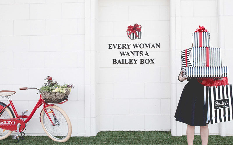 Person holding a striped box with 'Bailey's' branding next to a red bicycle with a basket of flowers against a white wall.