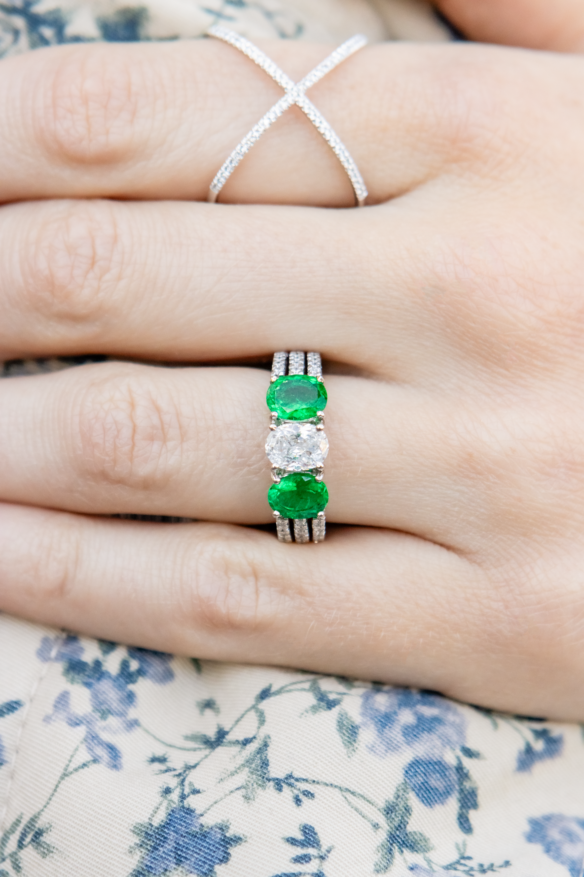 hand wearing diamond and emerald green rings on a floral blue and white fabric background