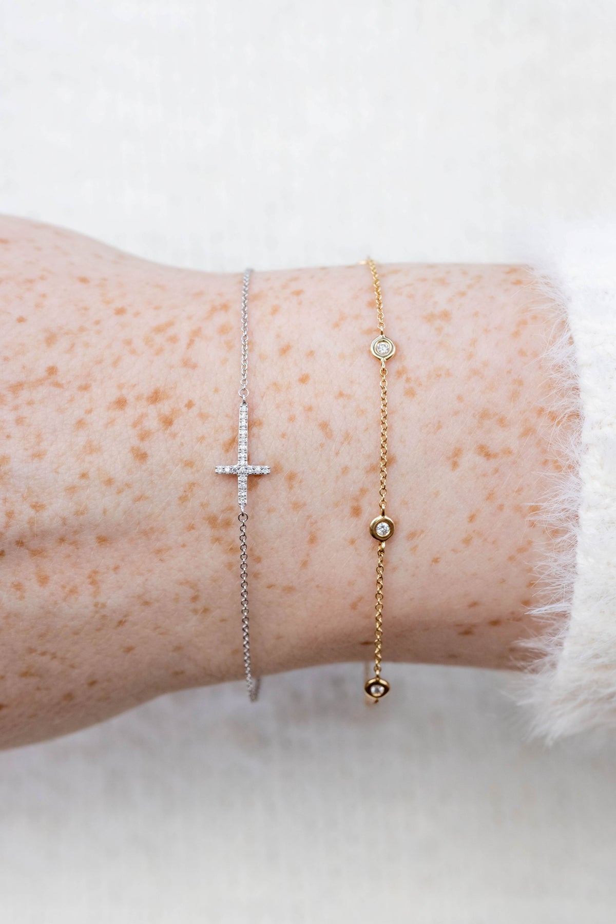 Two delicate bracelets worn on a person's wrist: a silver chain bracelet with a small cross and a gold chain bracelet with small bead details and a clear stone, shown against a soft white background.