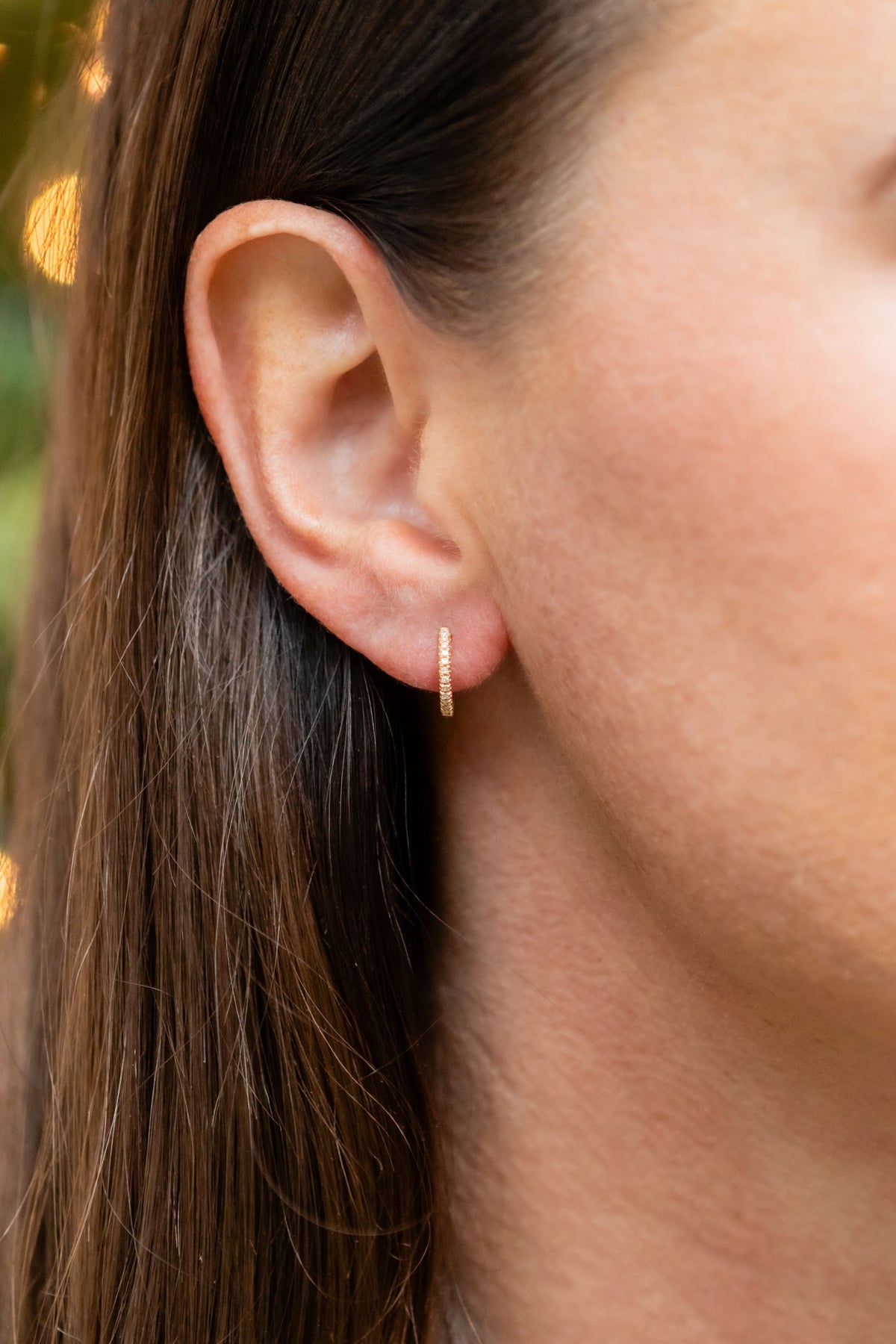 A close-up of a woman's ear wearing a delicate gold huggie hoop earring, with her hair and skin visible against a softly blurred outdoor background.