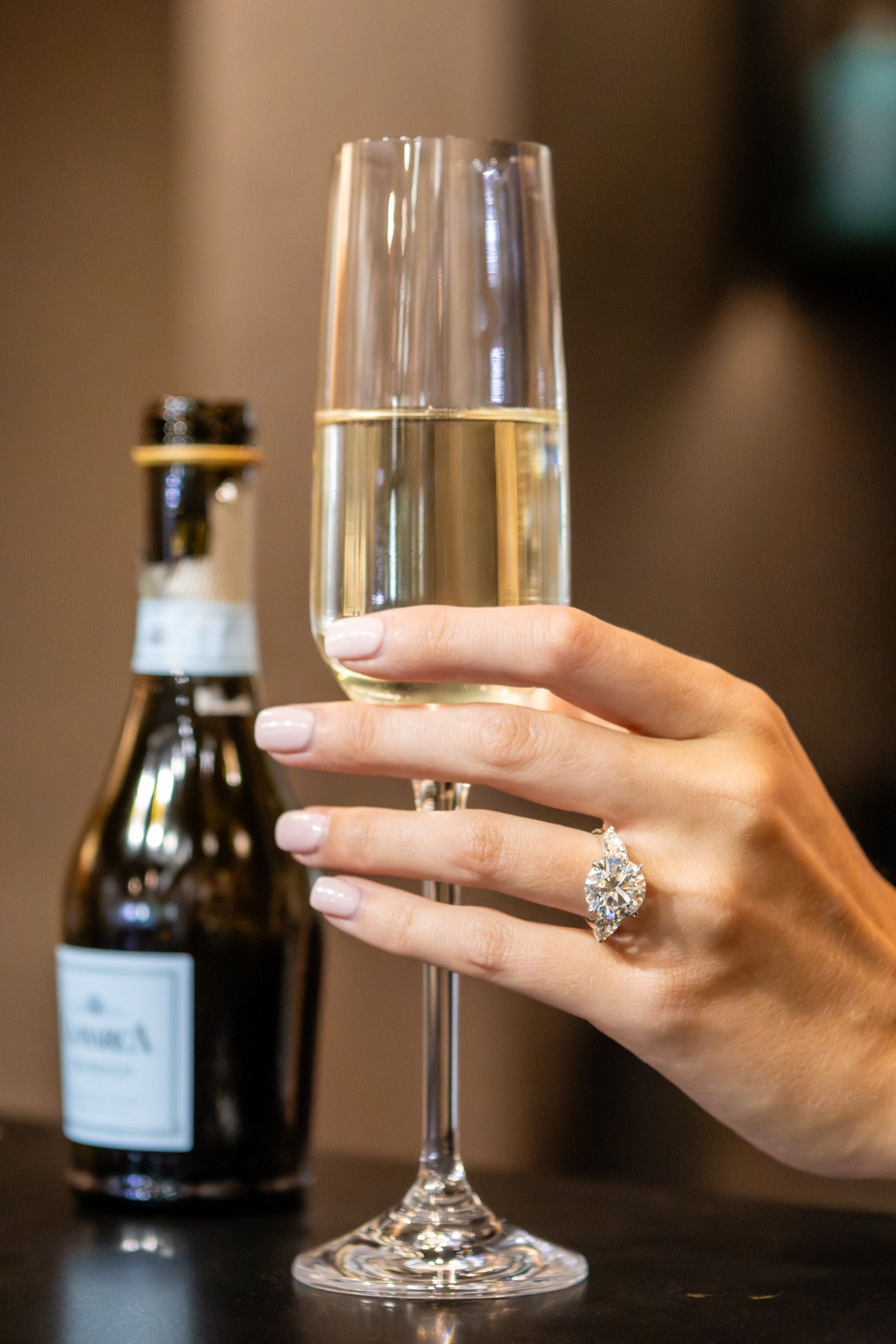 Hand with a diamond ring holding a glass of champagne next to a bottle on a table.