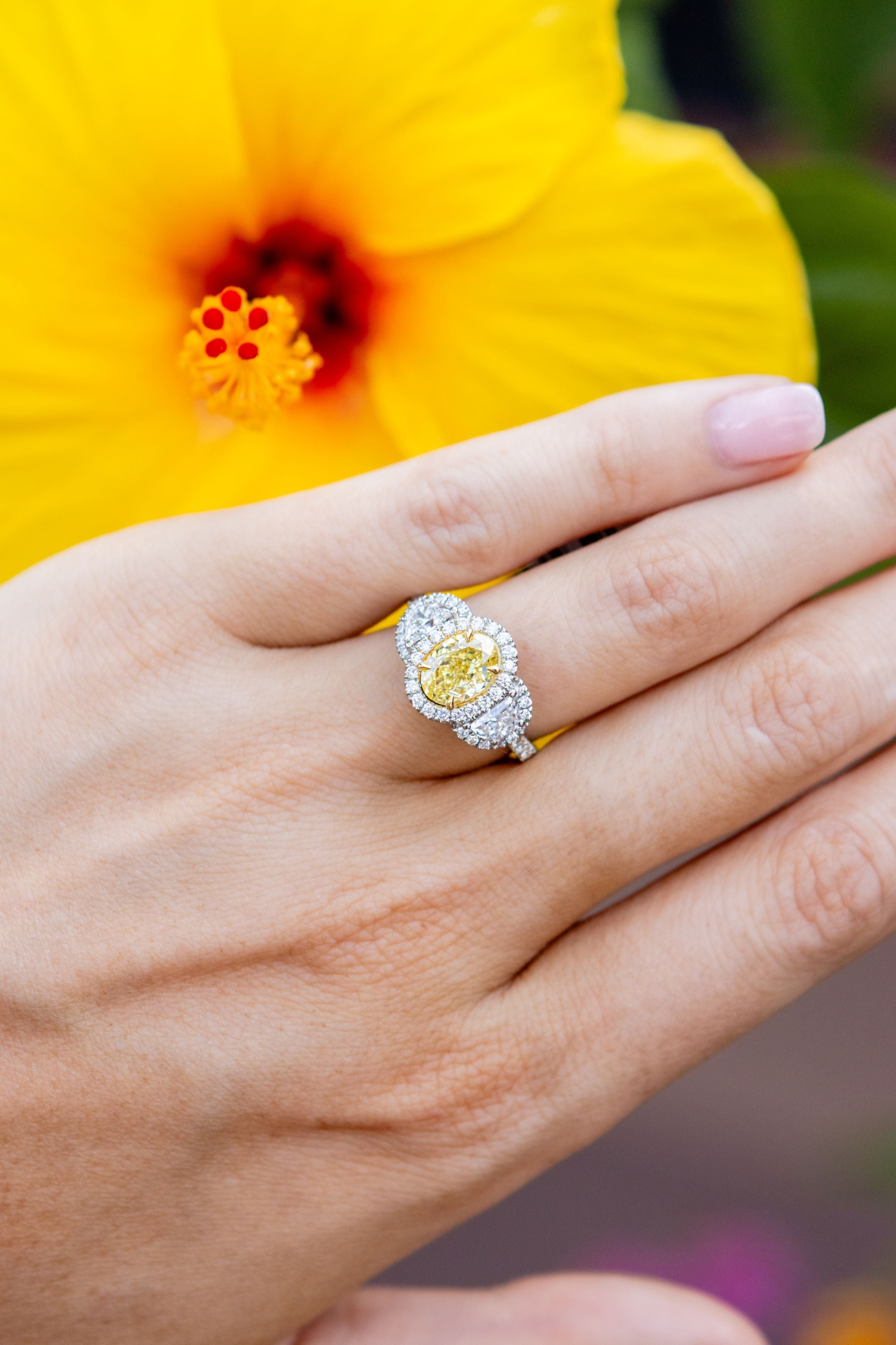 hand wearing yellow diamond ring in front of a large yellow flower with a blurred green background