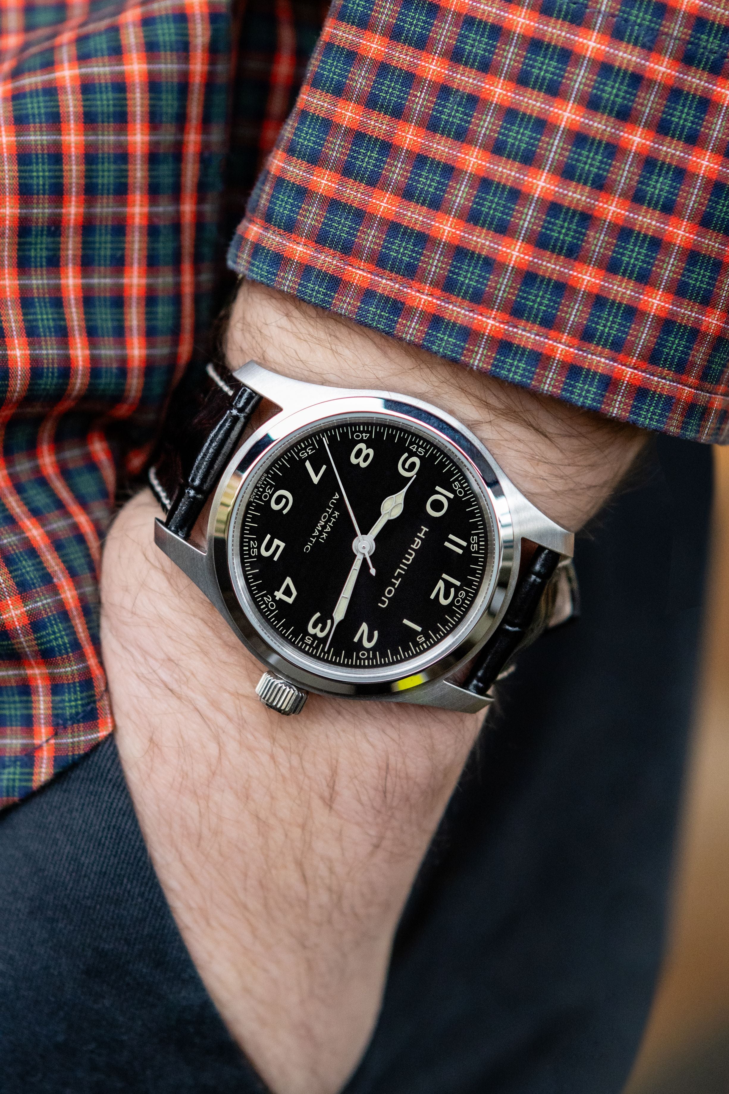 A close-up of a wrist wearing a Hamilton watch with a black dial and black leather strap, styled with a red, green, and blue plaid shirt cuff.
