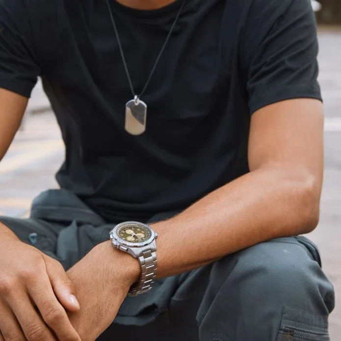 Person wearing a black t-shirt, gray pants, and a silver watch with a dog tag on a blurred background
