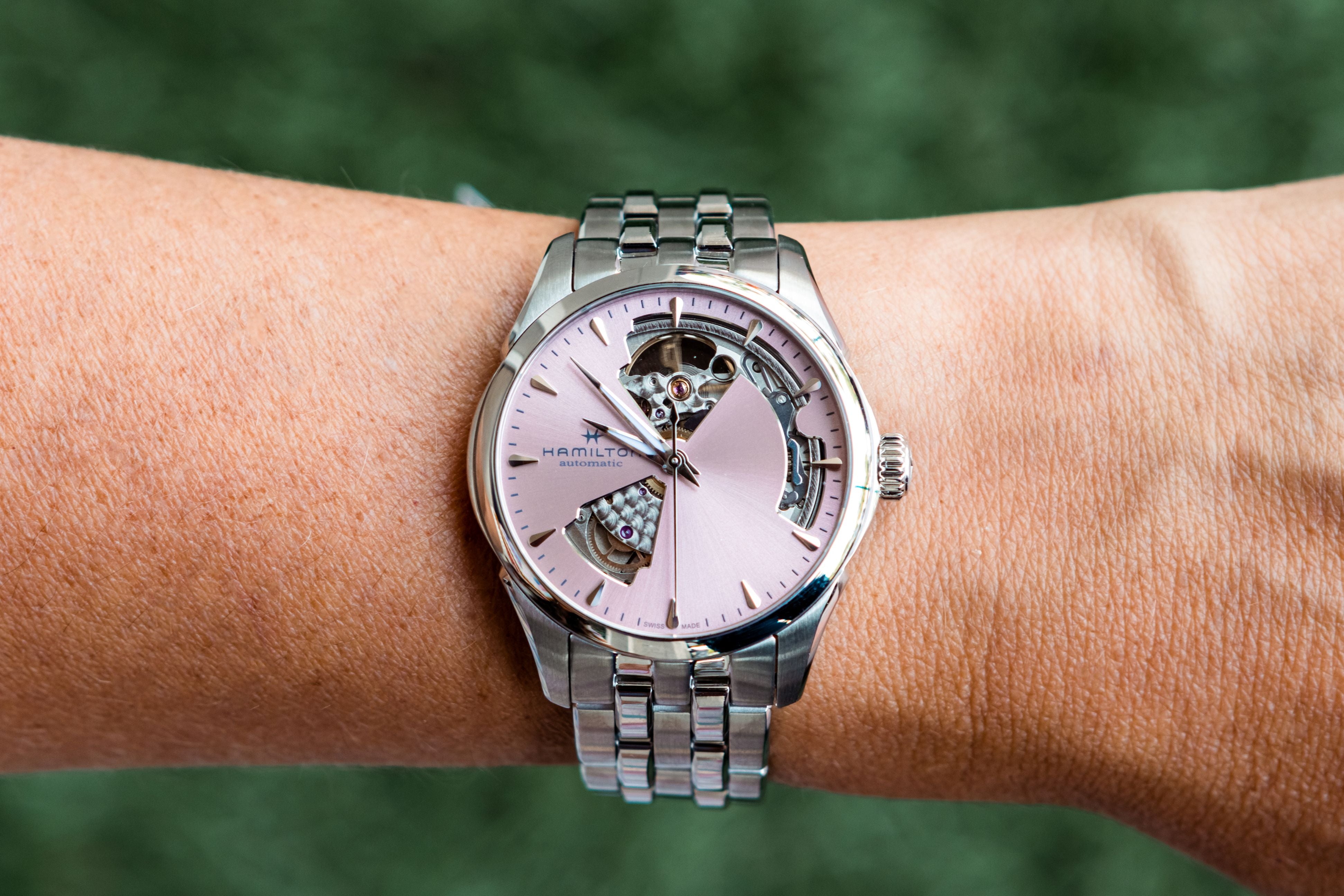A silver-tone wristwatch with a pink dial and skeletonized details, shown worn on a person's wrist against a blurred green background.