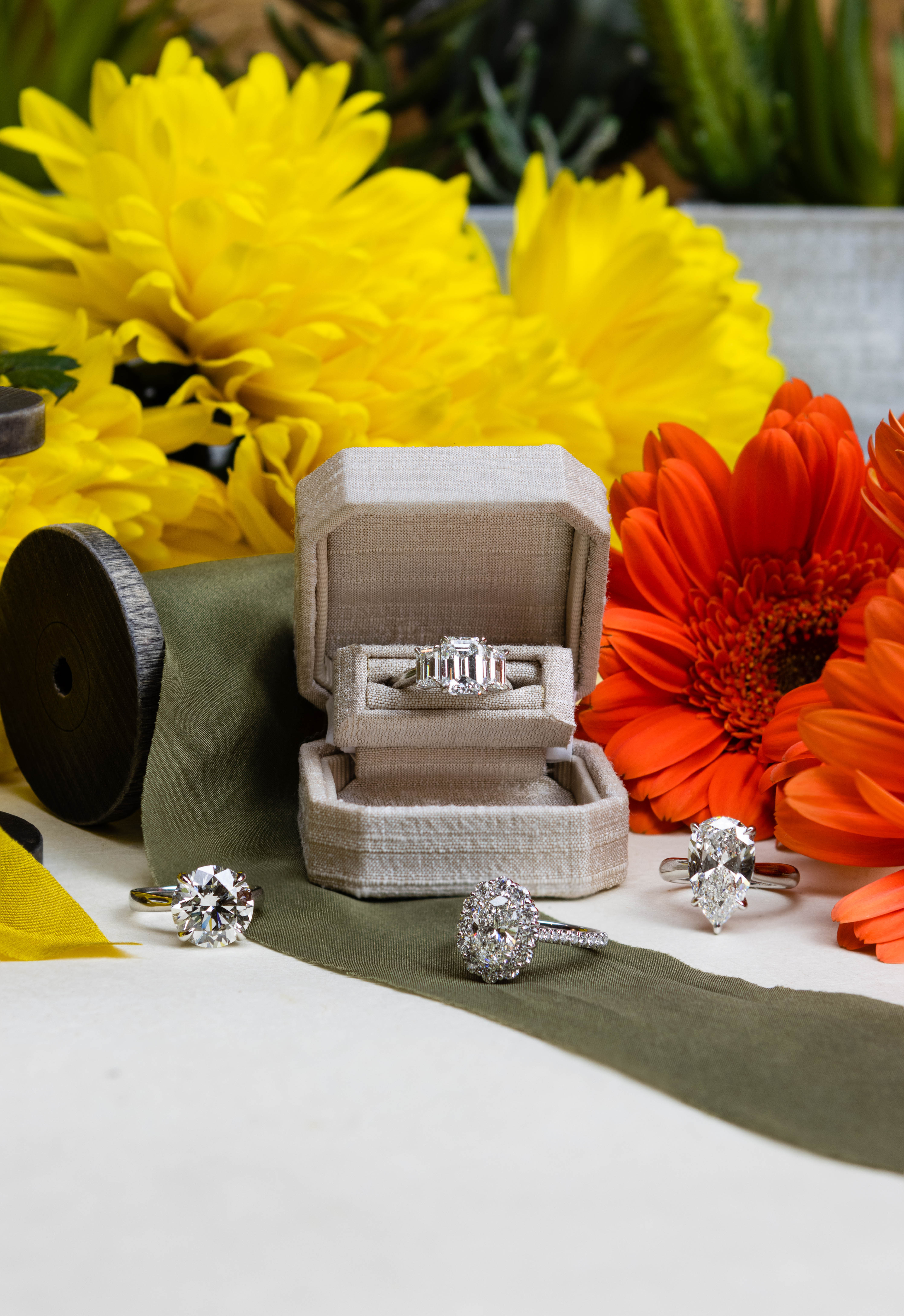 four silver diamond rings displayed around a beige box and yellow and red flowers on a white surface