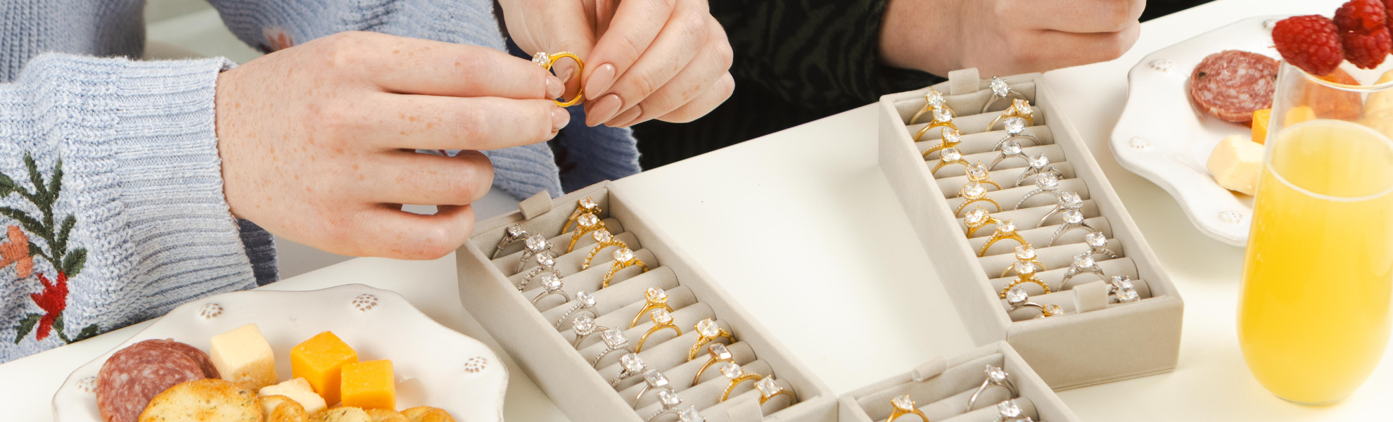 Person examining engagement rings in a jewelry box with snacks and drinks on a table.