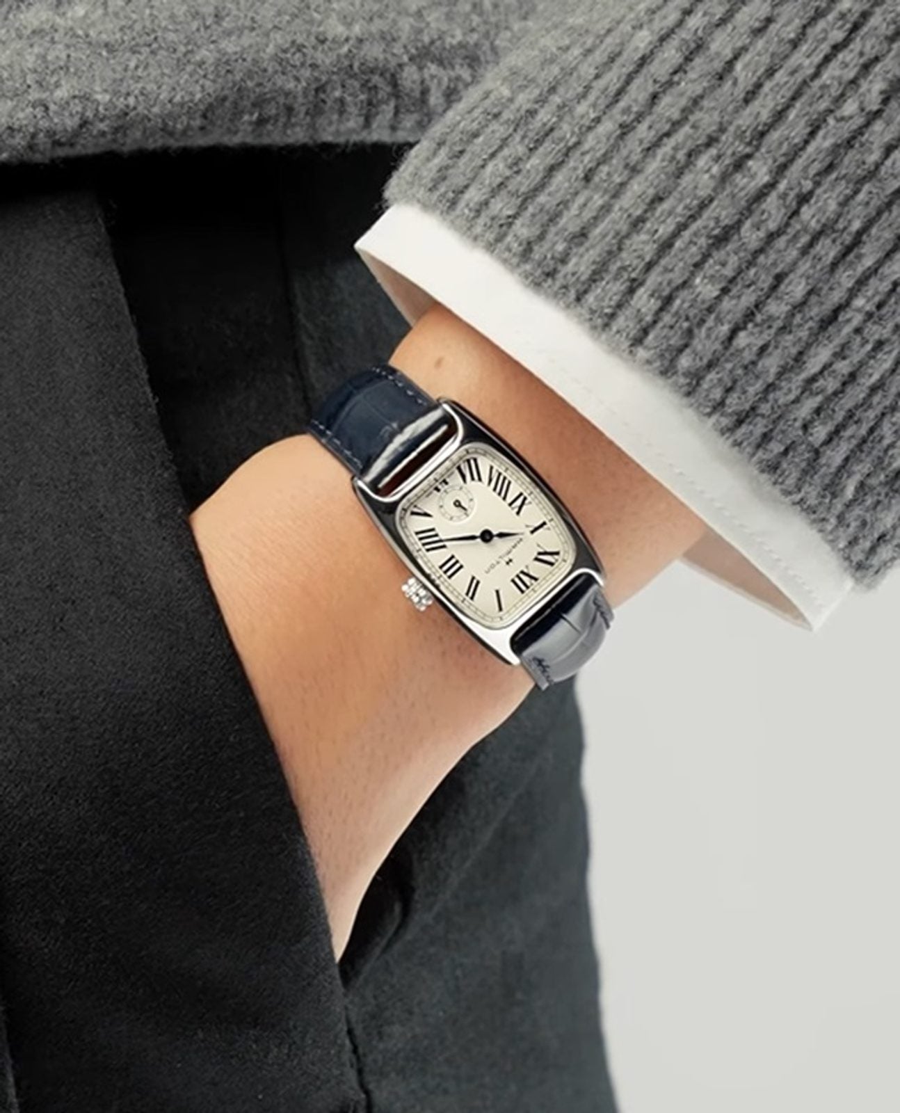 A close-up of a person's wrist wearing a slim rectangular watch with a silver-toned case, white dial, and dark navy blue leather strap, styled with a gray ribbed sweater and black blazer.
