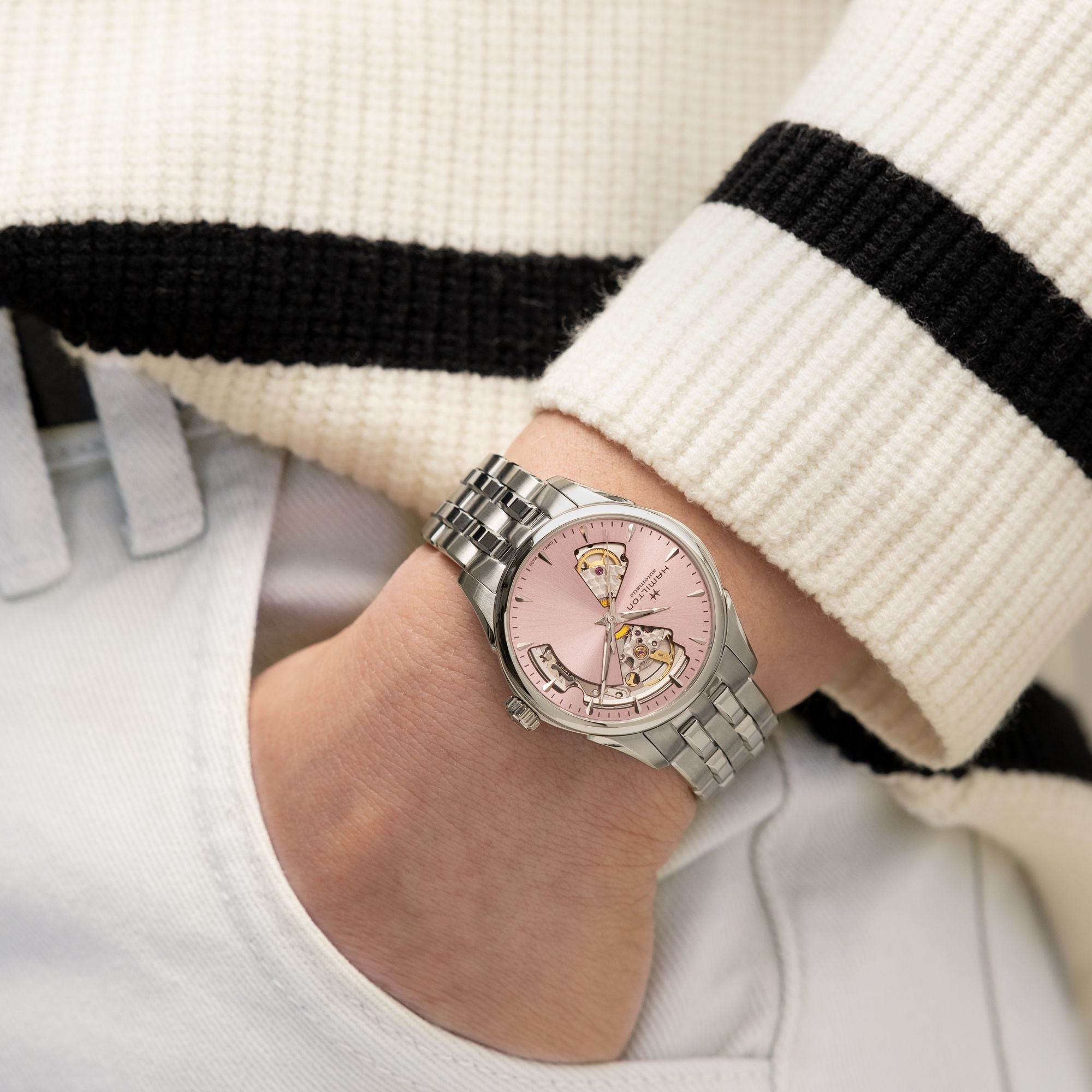 Hand wearing a silver watch with a pink face, against a blurred background.