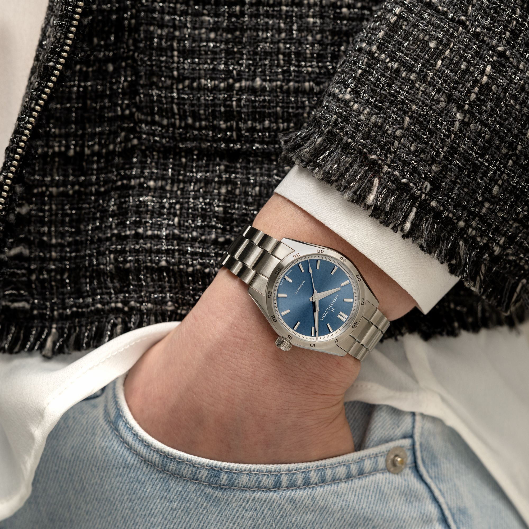 Silver watch with a blue face on a person's wrist, wearing a textured black jacket and denim jeans.