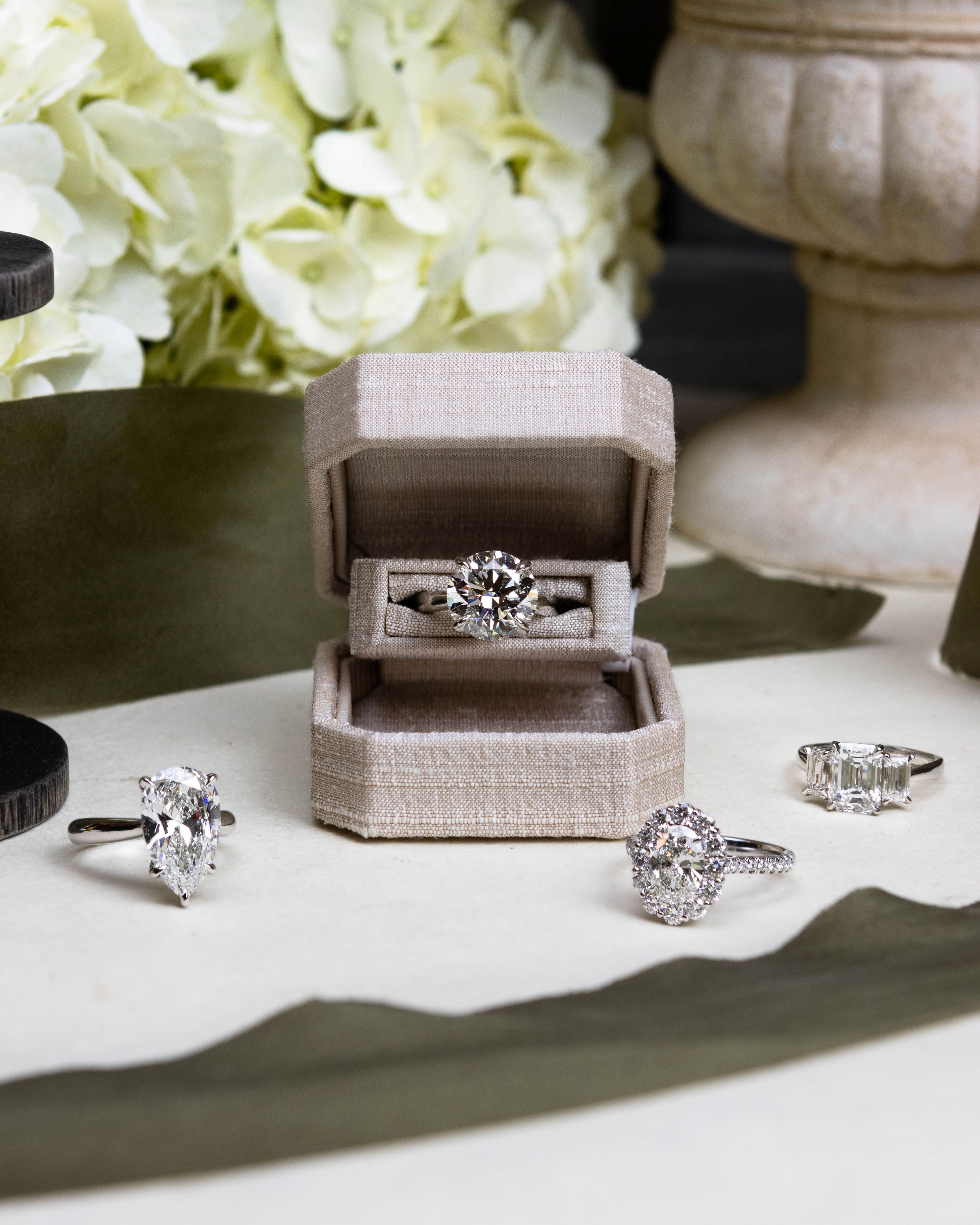 Diamond ring in an open jewelry box with flowers and other rings on a white surface