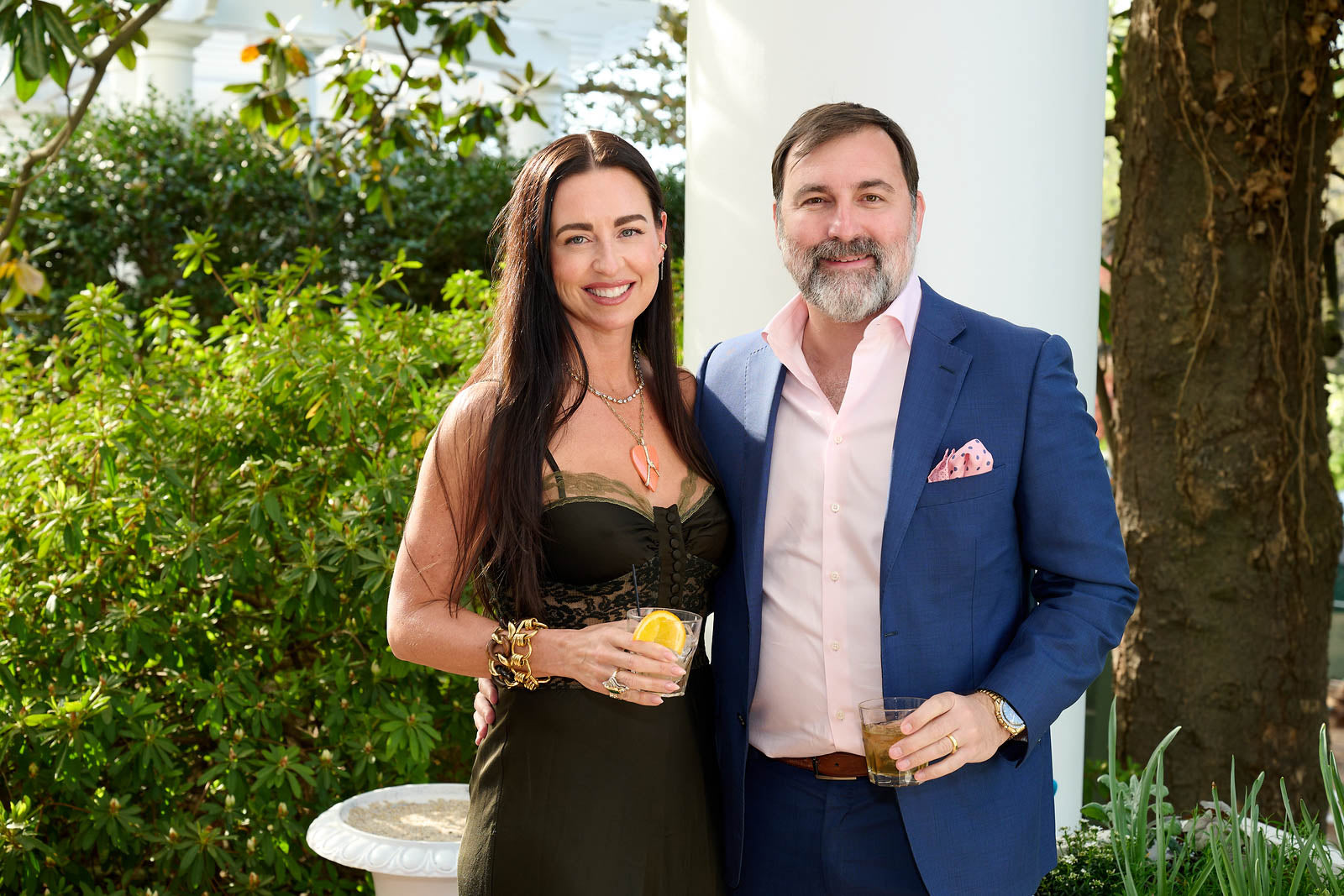 Man and woman standing together outdoors, holding drinks, with greenery in the background. Trey and Marci Bailey.