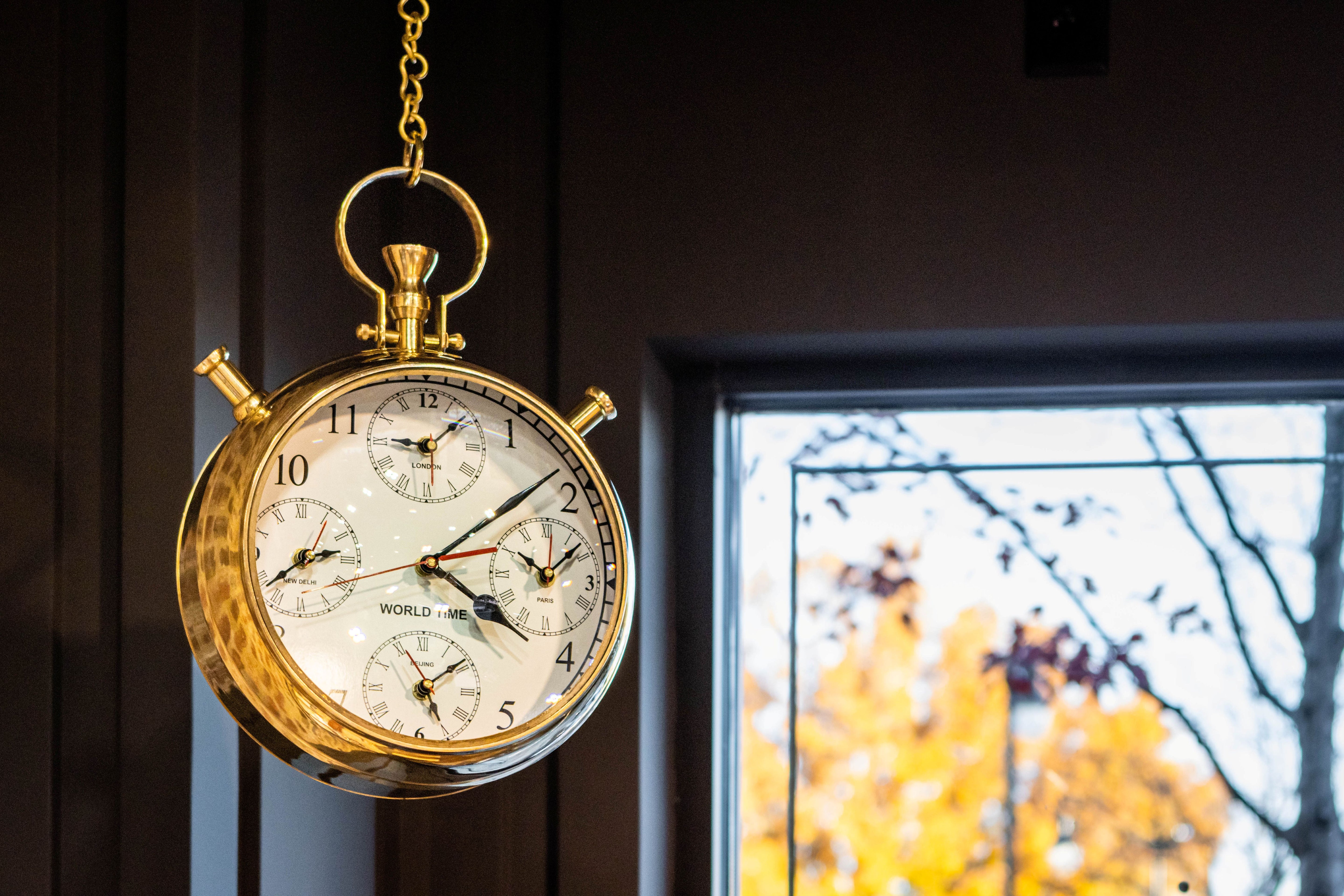 Gold pocket watch hanging against a dark background with a blurred window in the background.