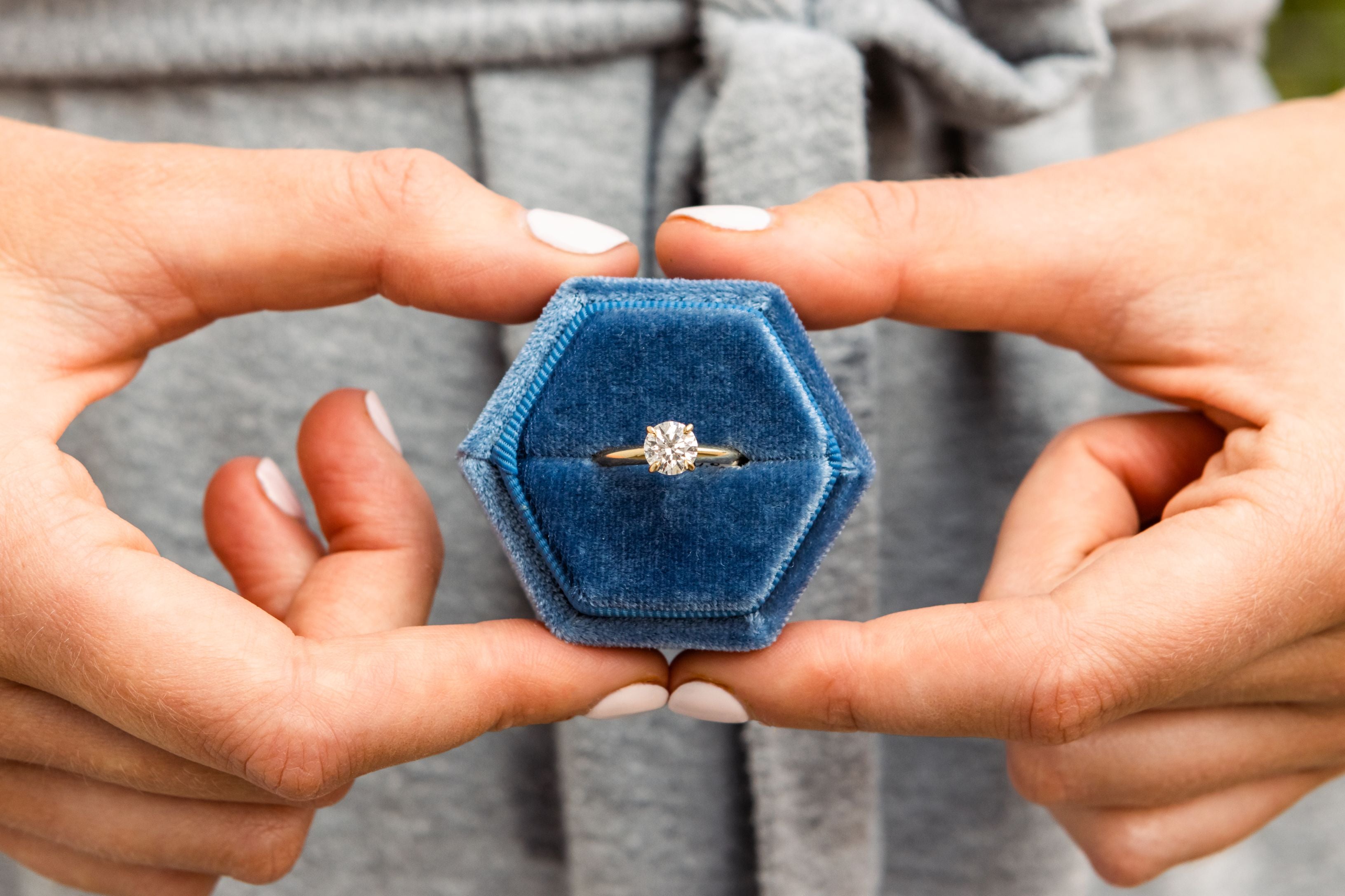 two hands holding a diamond ring in a blue hexagonal box with a blurred grey background