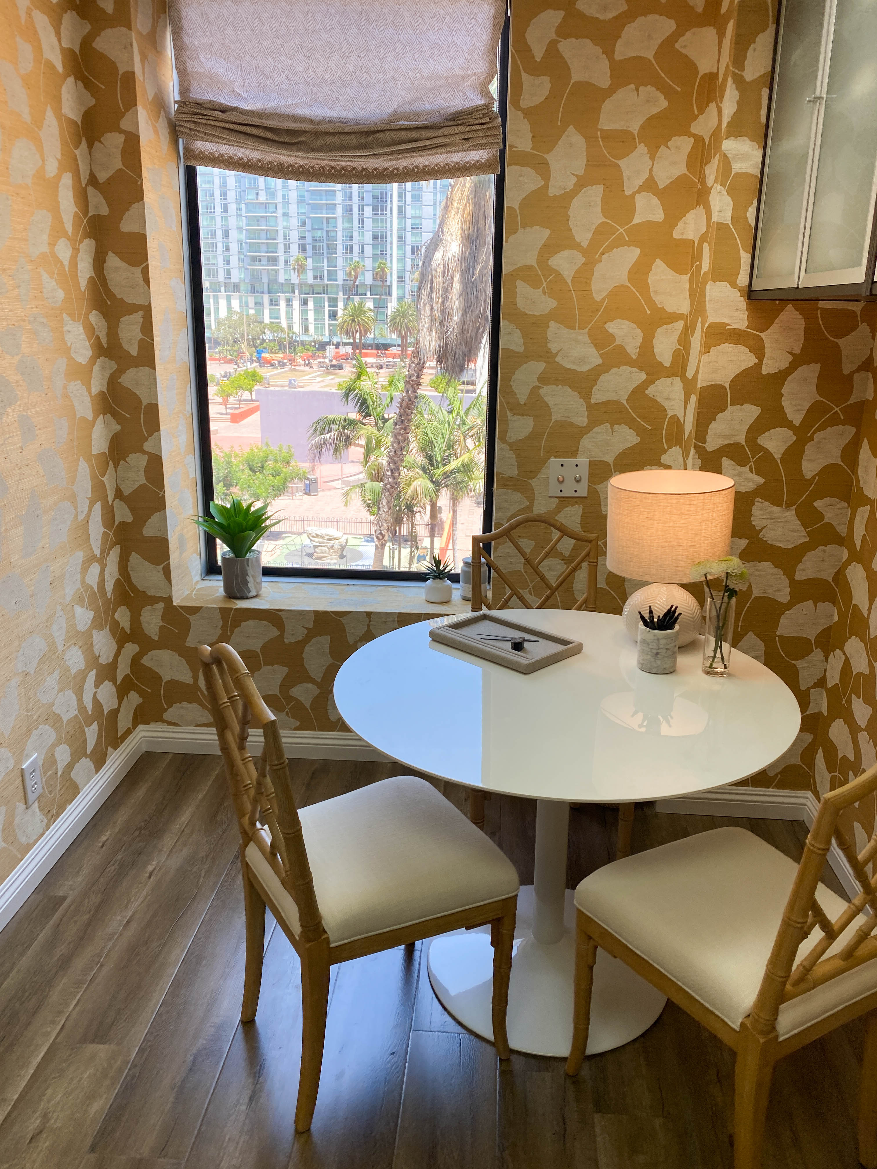 Dining area with round table and chairs in a room with patterned walls and a view of buildings and palm trees outside.