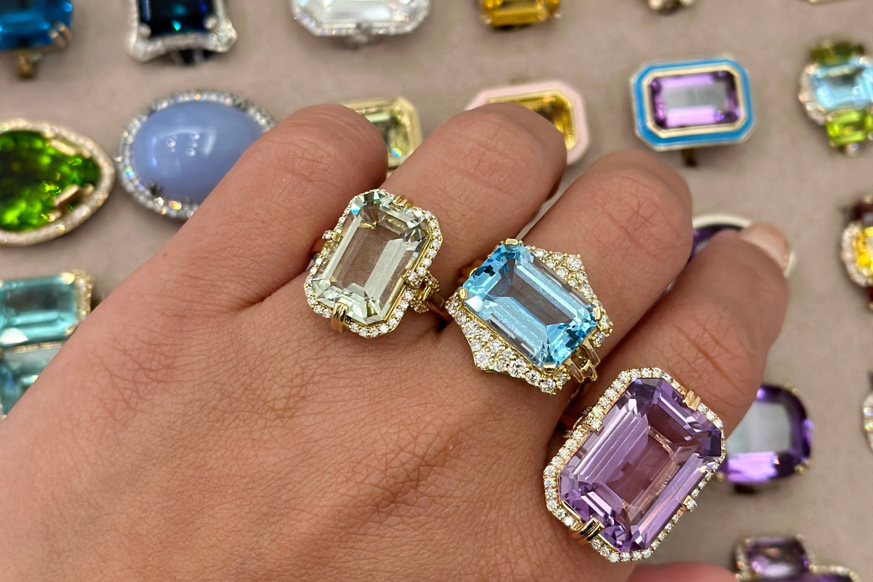 Hand wearing two gemstone rings with a background of various gemstones.