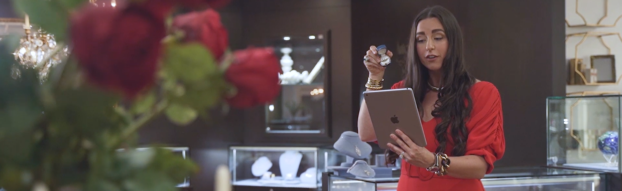 Woman in a red dress holding a tablet in a jewelry store with red roses in the foreground.