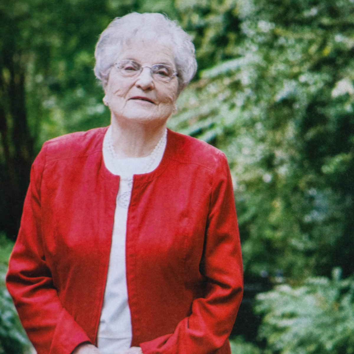Woman wearing a red jacket and glasses standing in front of green foliage