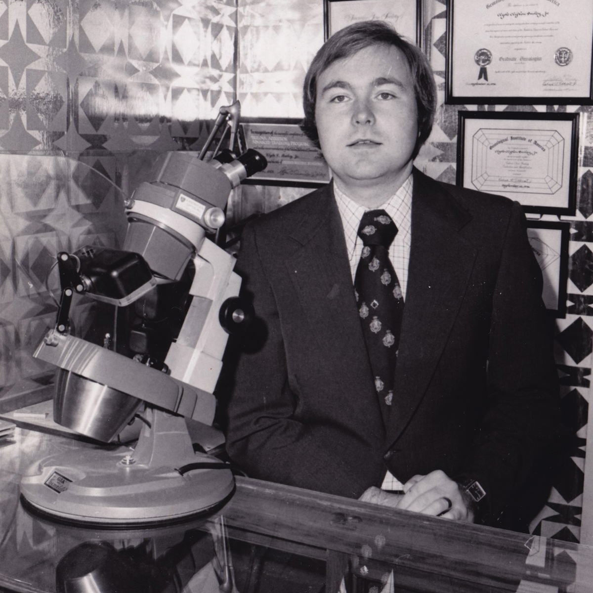 Man in a suit sitting next to a microscope in an office setting with framed documents on the wall.